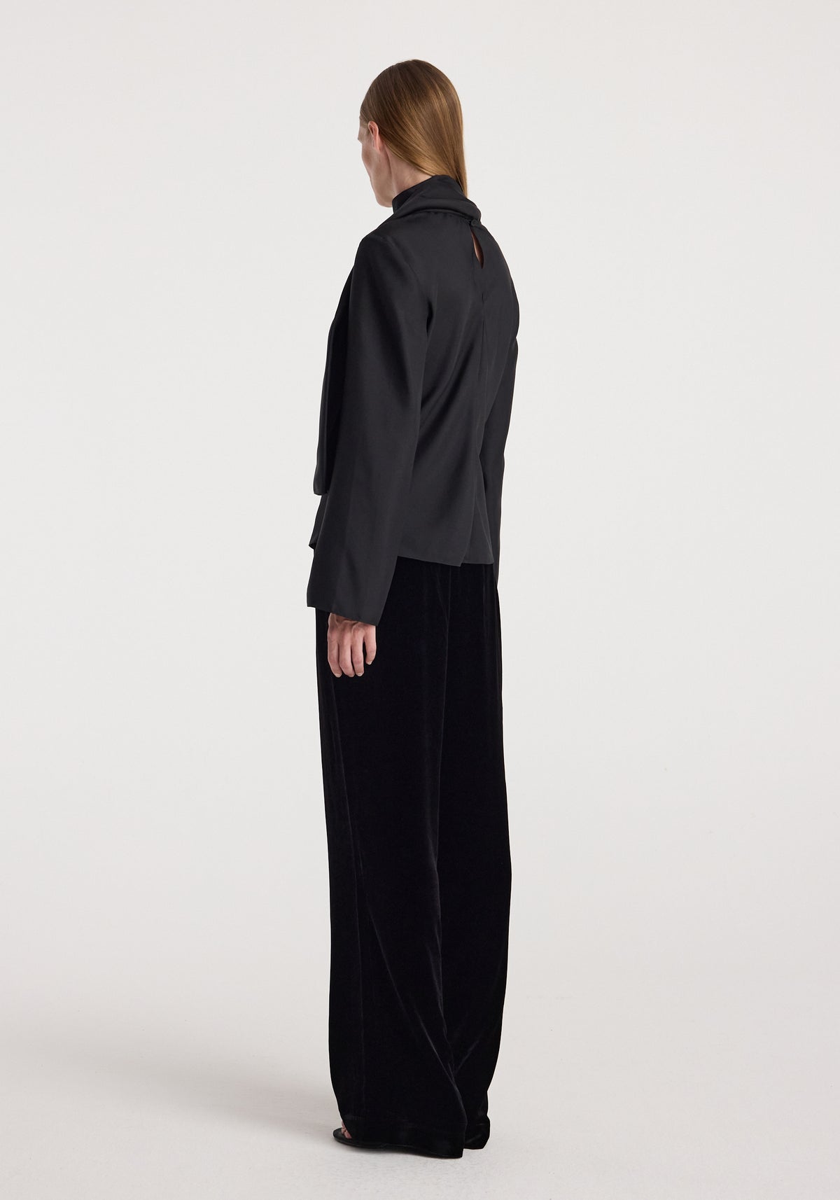 Woman wearing Velvet relaxed trousers | black with a high-waisted, loose fit and wide legs, paired with a matching black long-sleeve top against a plain background.