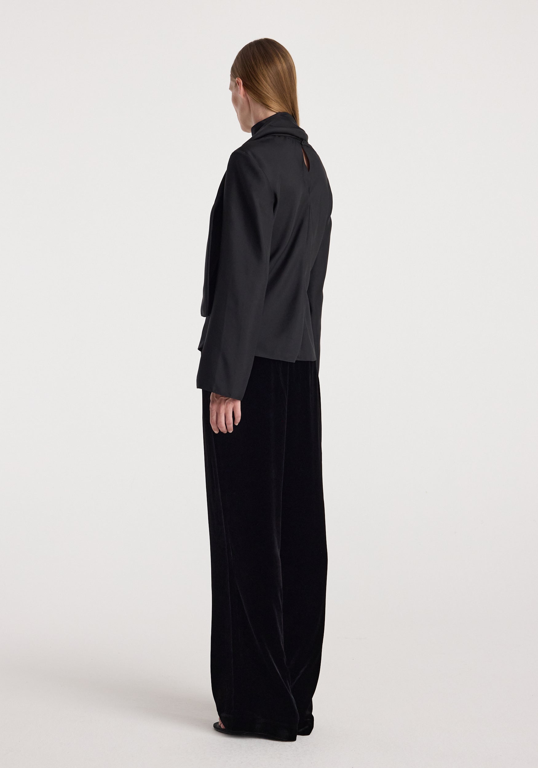 Woman wearing Velvet relaxed trousers | black with a high-waisted, loose fit and wide legs, paired with a matching black long-sleeve top against a plain background.