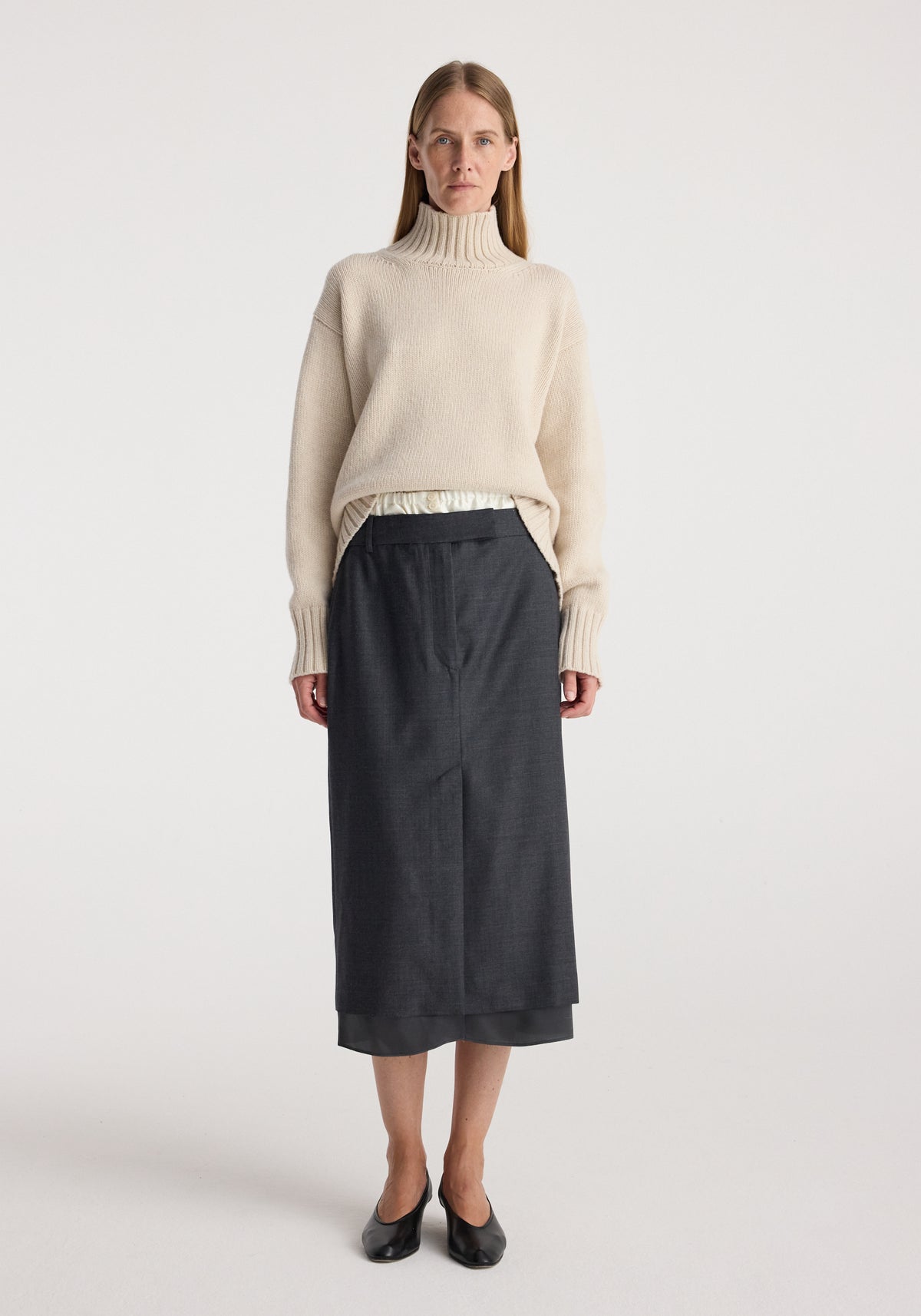 Woman wearing a cream turtleneck sweater paired with a Boxer detailed skirt | dark grey melange, standing against a plain white background with black flats.