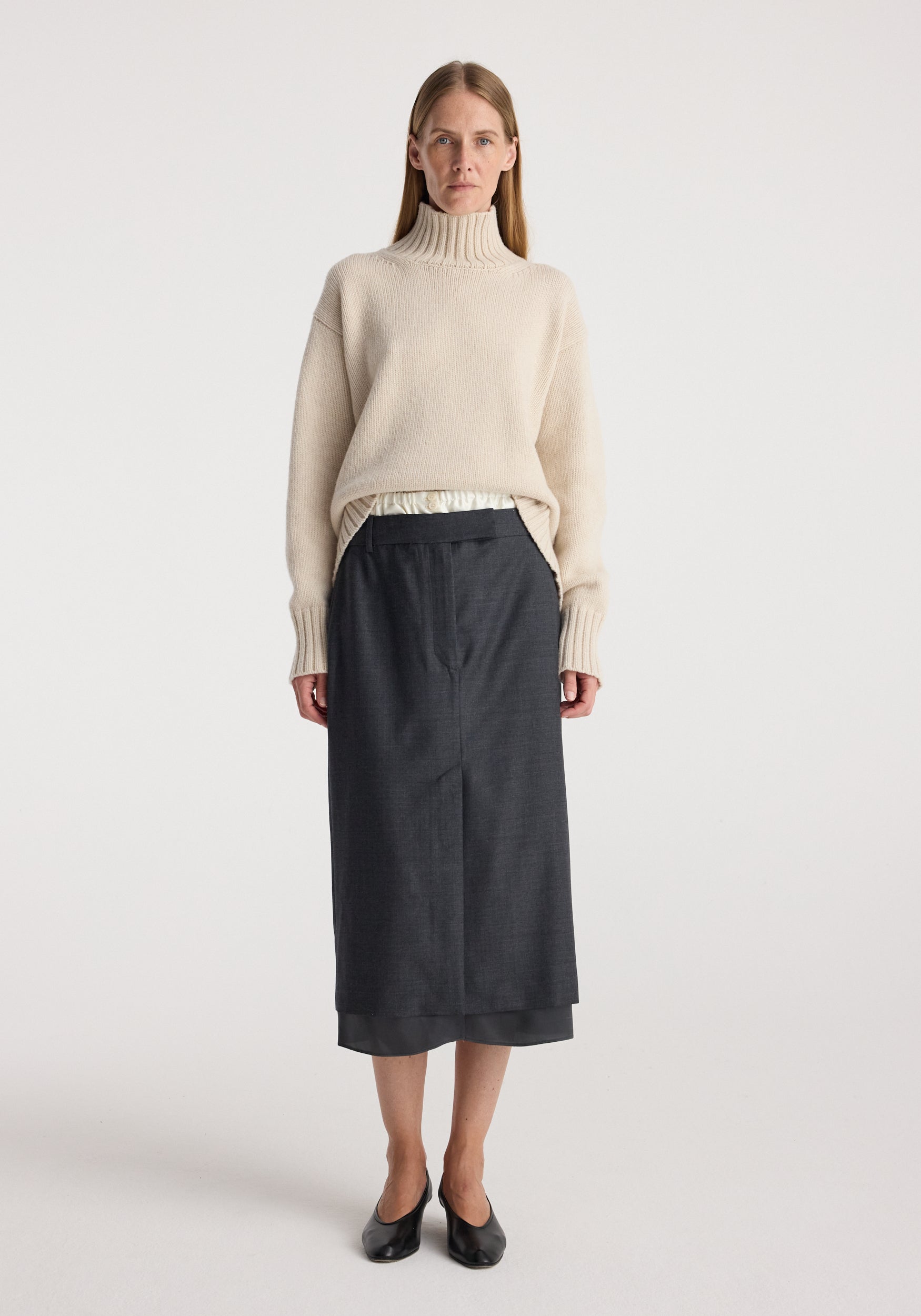 Woman wearing a cream turtleneck sweater paired with a Boxer detailed skirt | dark grey melange, standing against a plain white background with black flats.