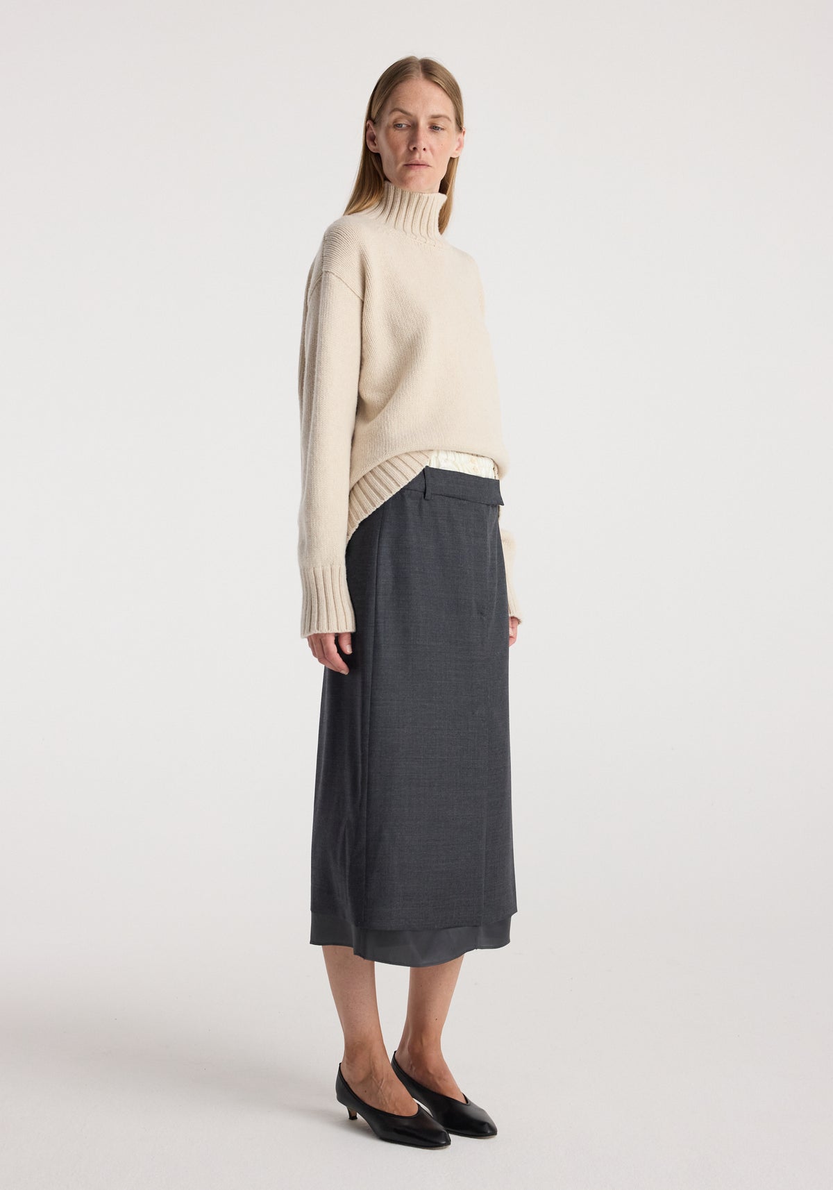 Woman wearing a beige turtleneck sweater paired with a dark grey melange boxer detailed skirt | dark grey melange and black low-heeled shoes, standing against a plain background.