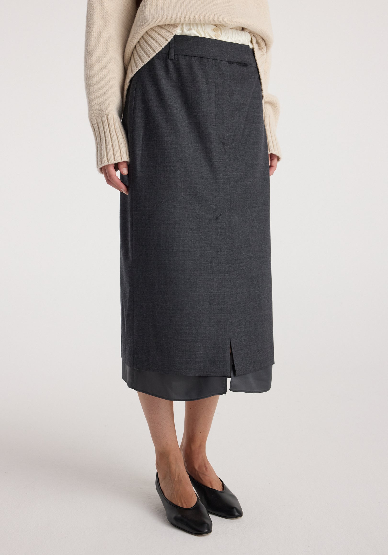 Woman wearing a dark grey melange skirt layered over a satin underskirt and paired with a beige sweater and black pointed flats, showcasing the Boxer detailed skirt | dark grey melange.