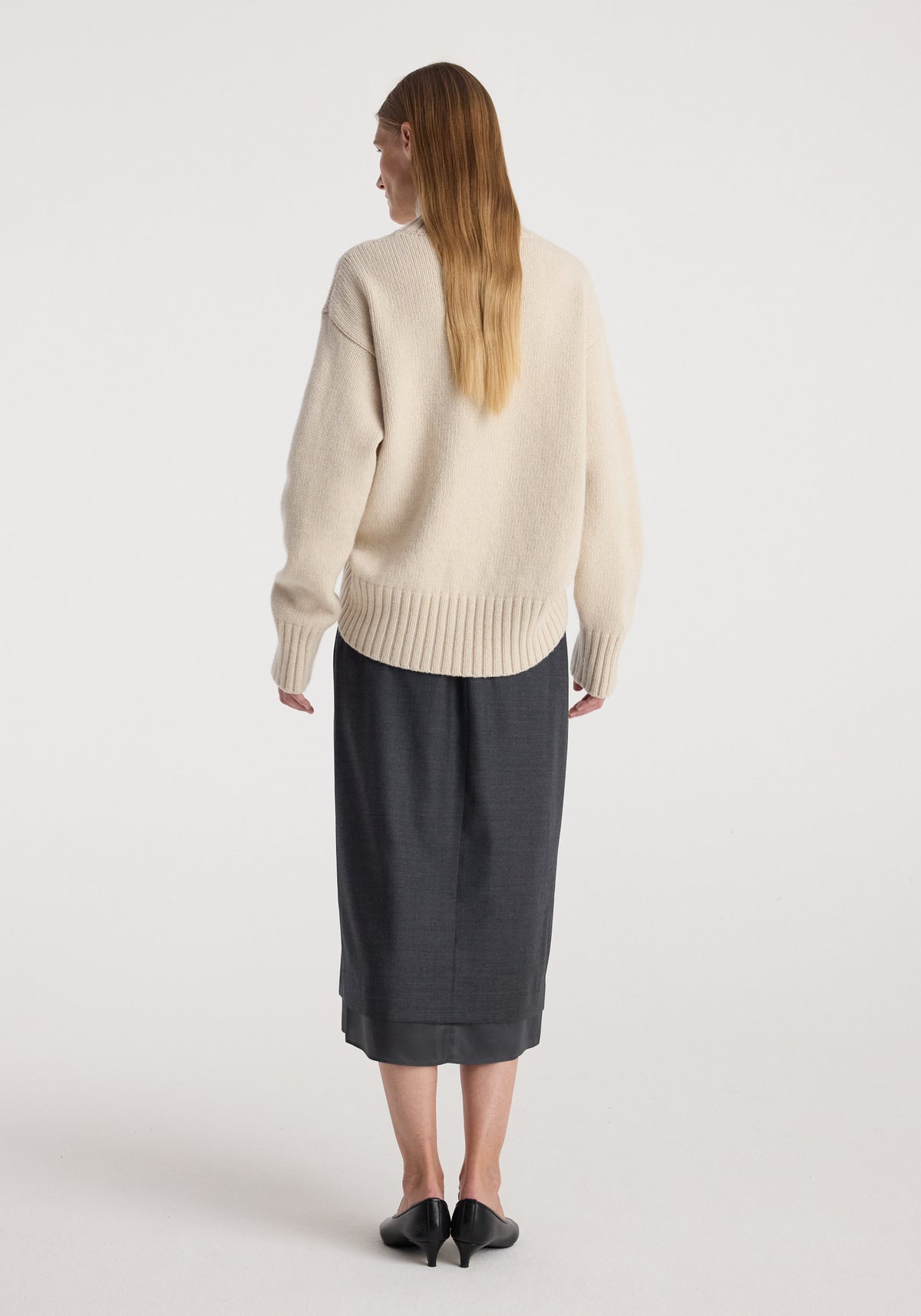 Woman seen from the back wearing a cream-colored knit sweater paired with a dark grey melange Boxer detailed skirt | dark grey melange and black low-heeled shoes, standing against a plain white background.