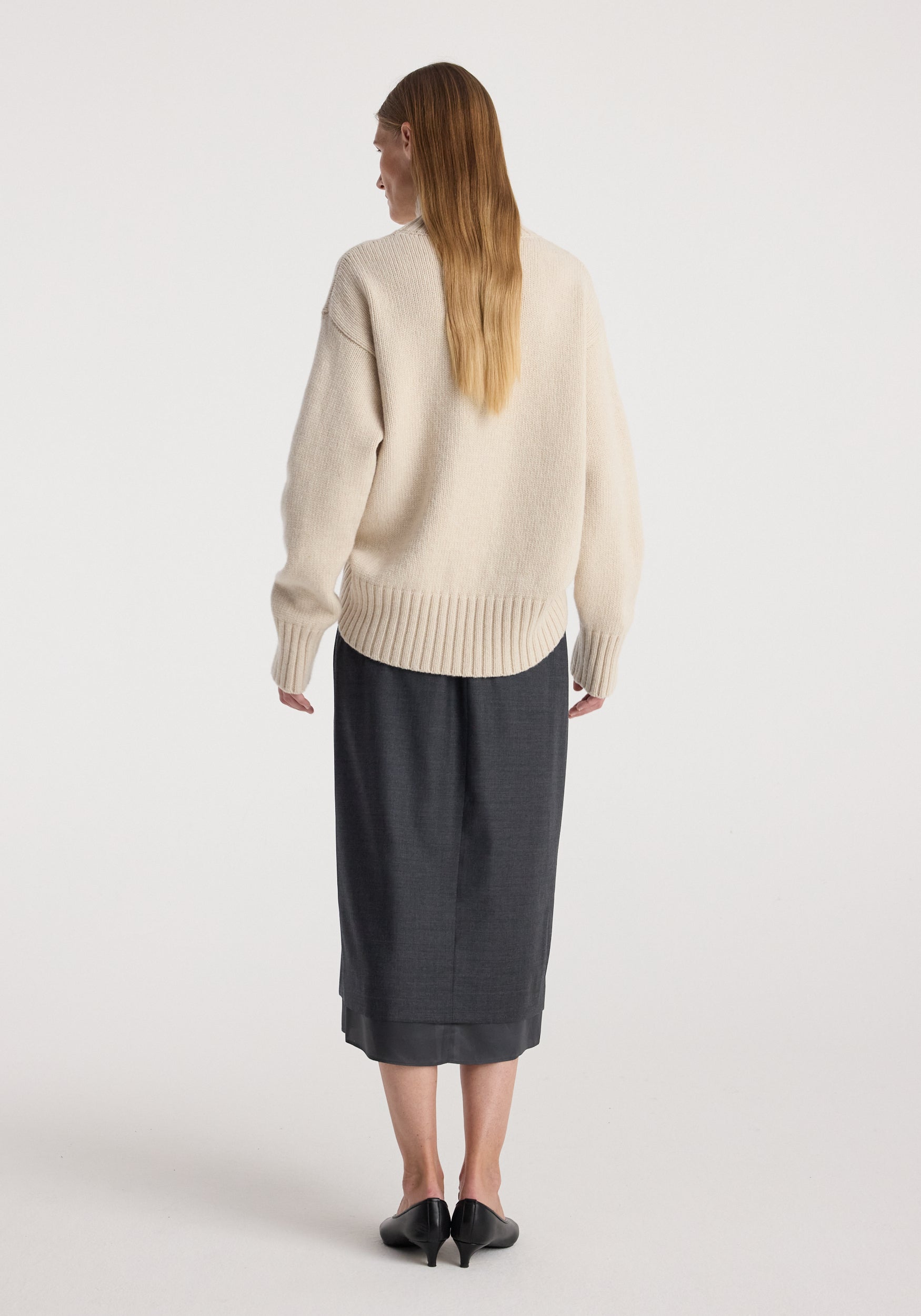 Woman seen from the back wearing a cream-colored knit sweater paired with a dark grey melange Boxer detailed skirt | dark grey melange and black low-heeled shoes, standing against a plain white background.