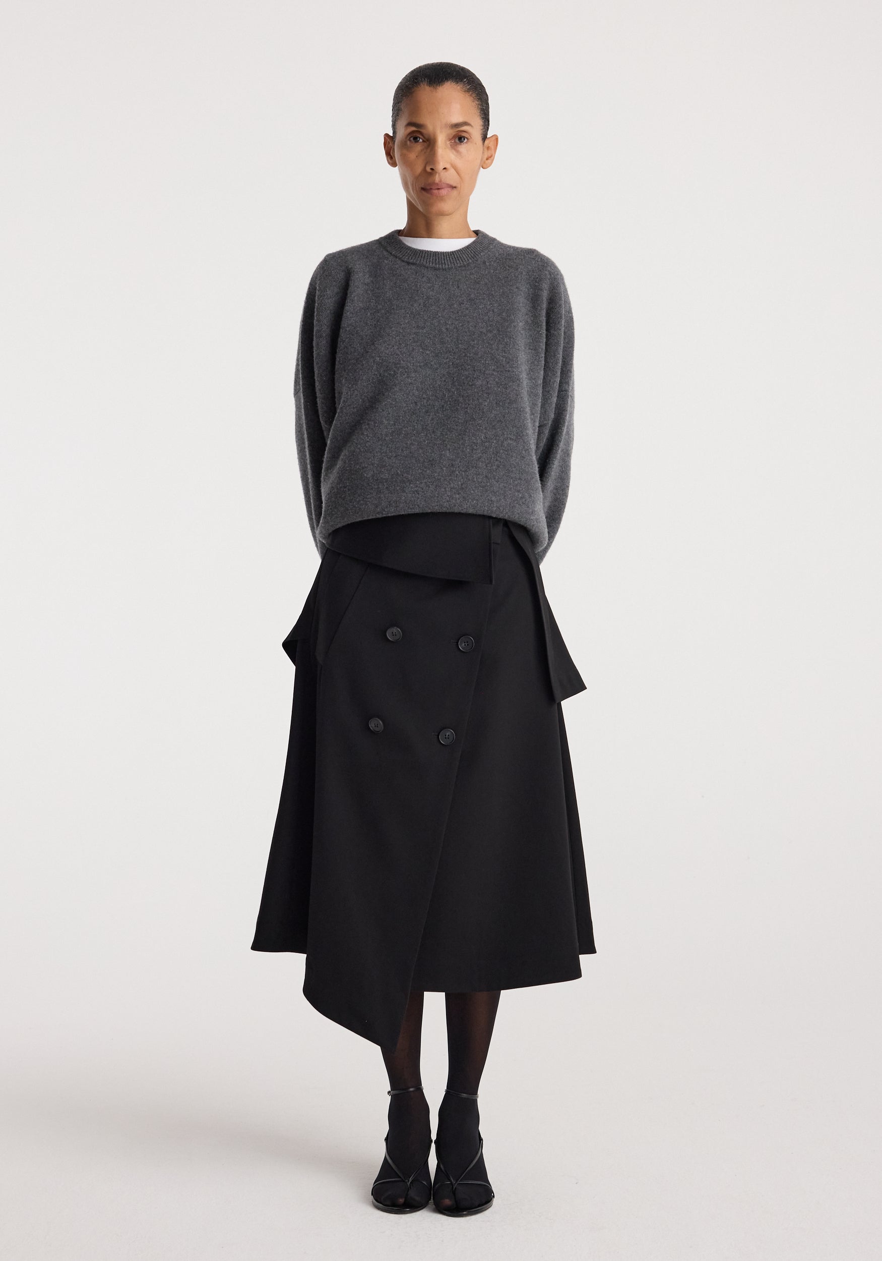 Woman wearing a gray sweater paired with a black asymmetrical buttoned skirt featuring flap details, styled with black tights and black open-toe shoes, showcasing the Deconstructed trench skirt | black.