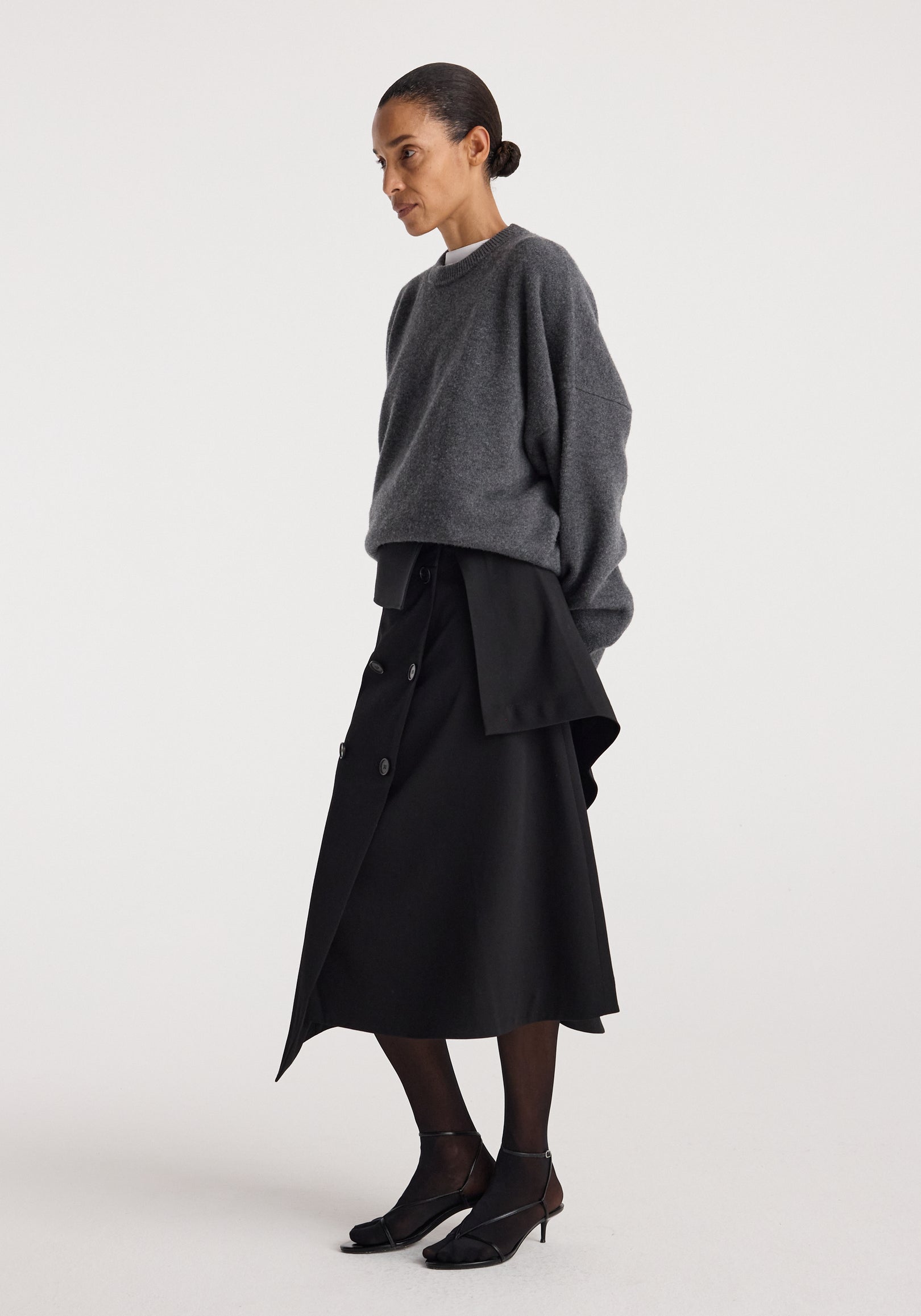 Woman wearing a dark gray oversized sweater paired with a black asymmetrical Deconstructed trench skirt | black, sheer black tights, and black heeled sandals against a plain background.