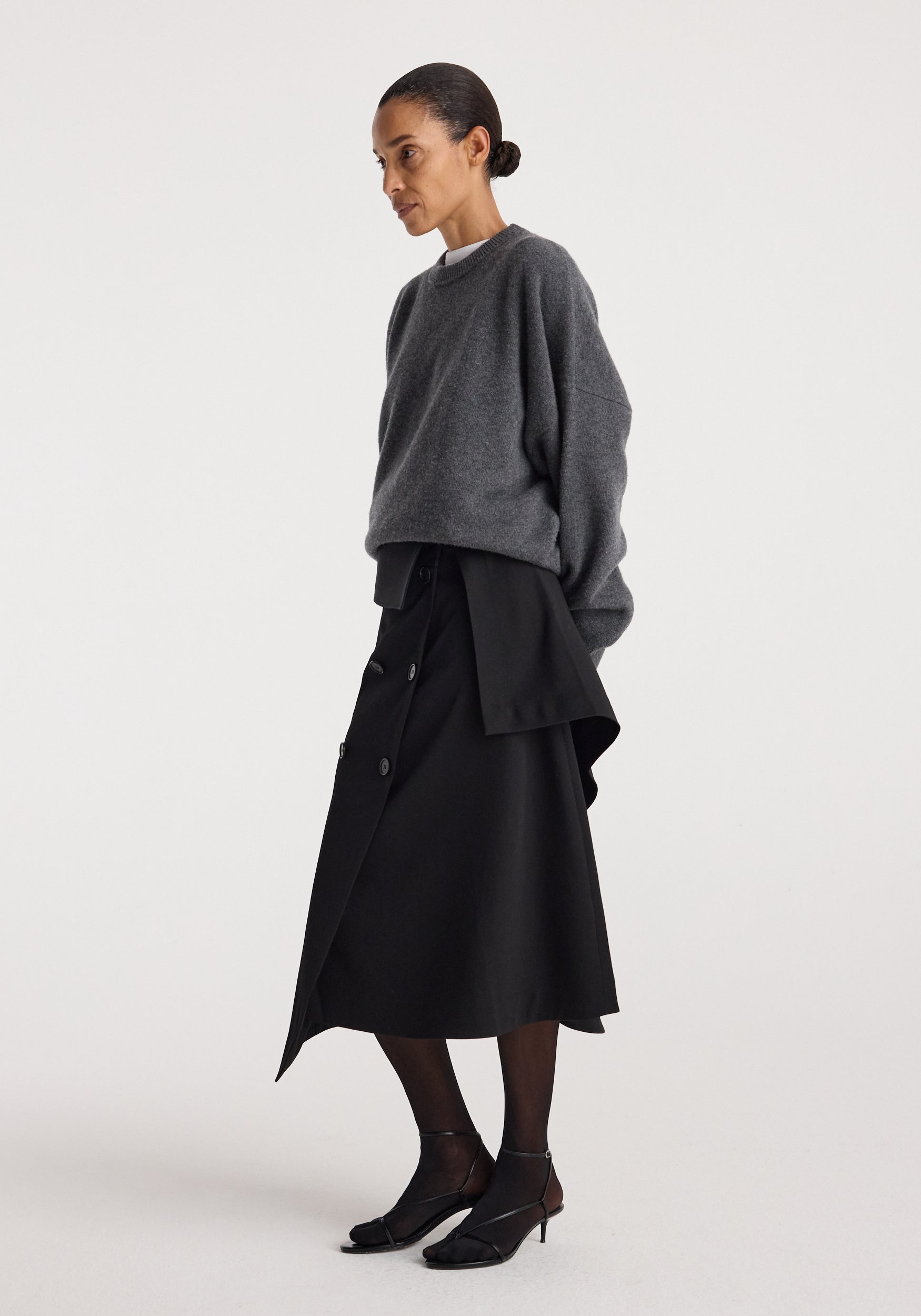 Woman wearing a dark gray oversized sweater paired with a black asymmetrical Deconstructed trench skirt | black, sheer black tights, and black heeled sandals against a plain background.