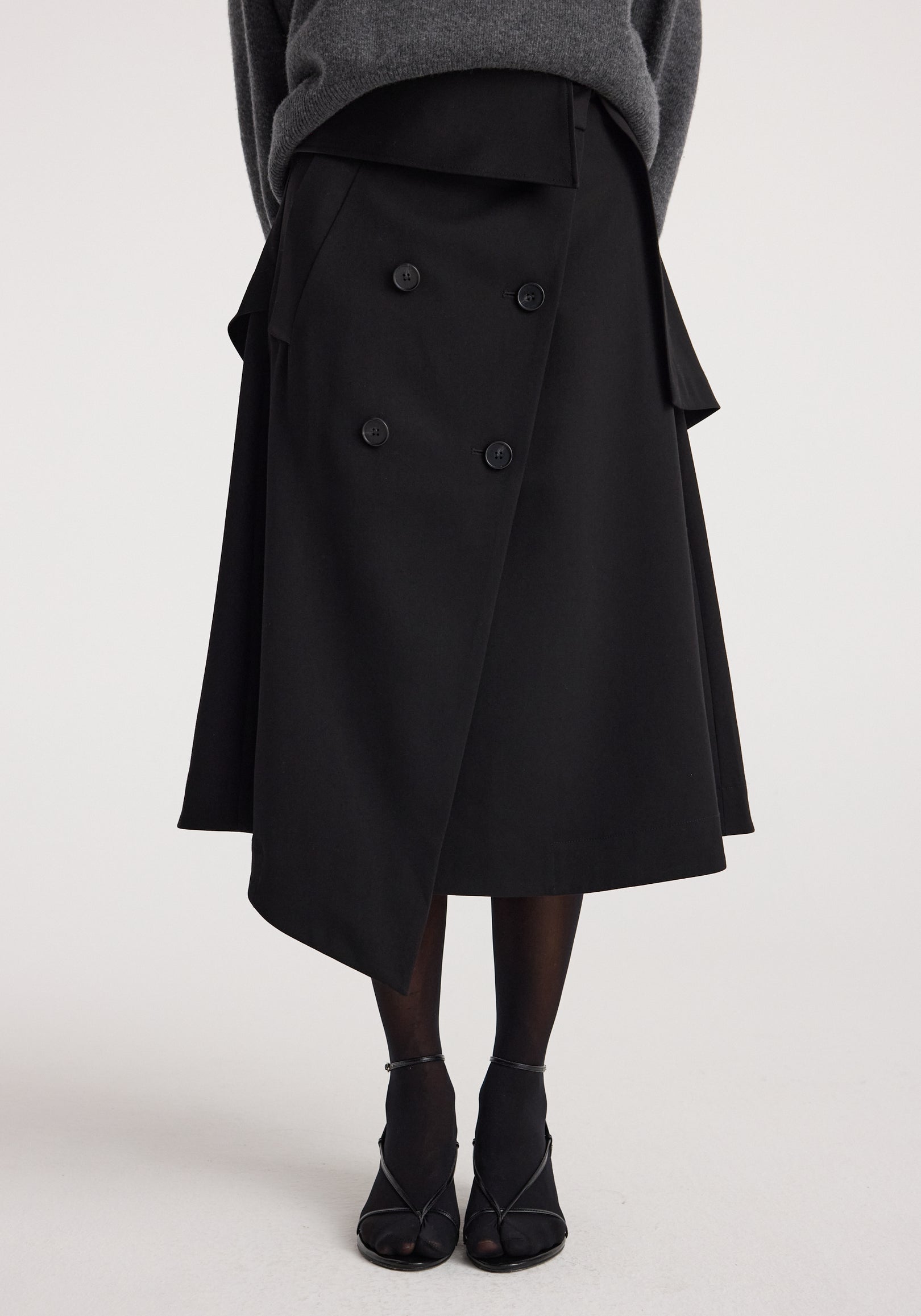 Front view of a person wearing a black asymmetrical skirt with double-breasted button details and an overlapping design, paired with black tights and heeled sandals, showcasing the Deconstructed trench skirt | black.