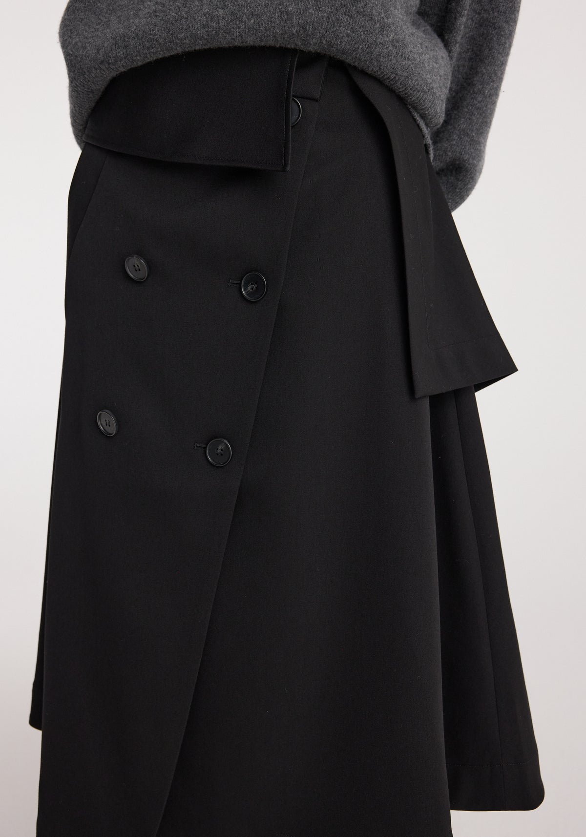 Close-up of a black Deconstructed trench skirt | black featuring button details and an asymmetrical layered design paired with a dark grey sweater.