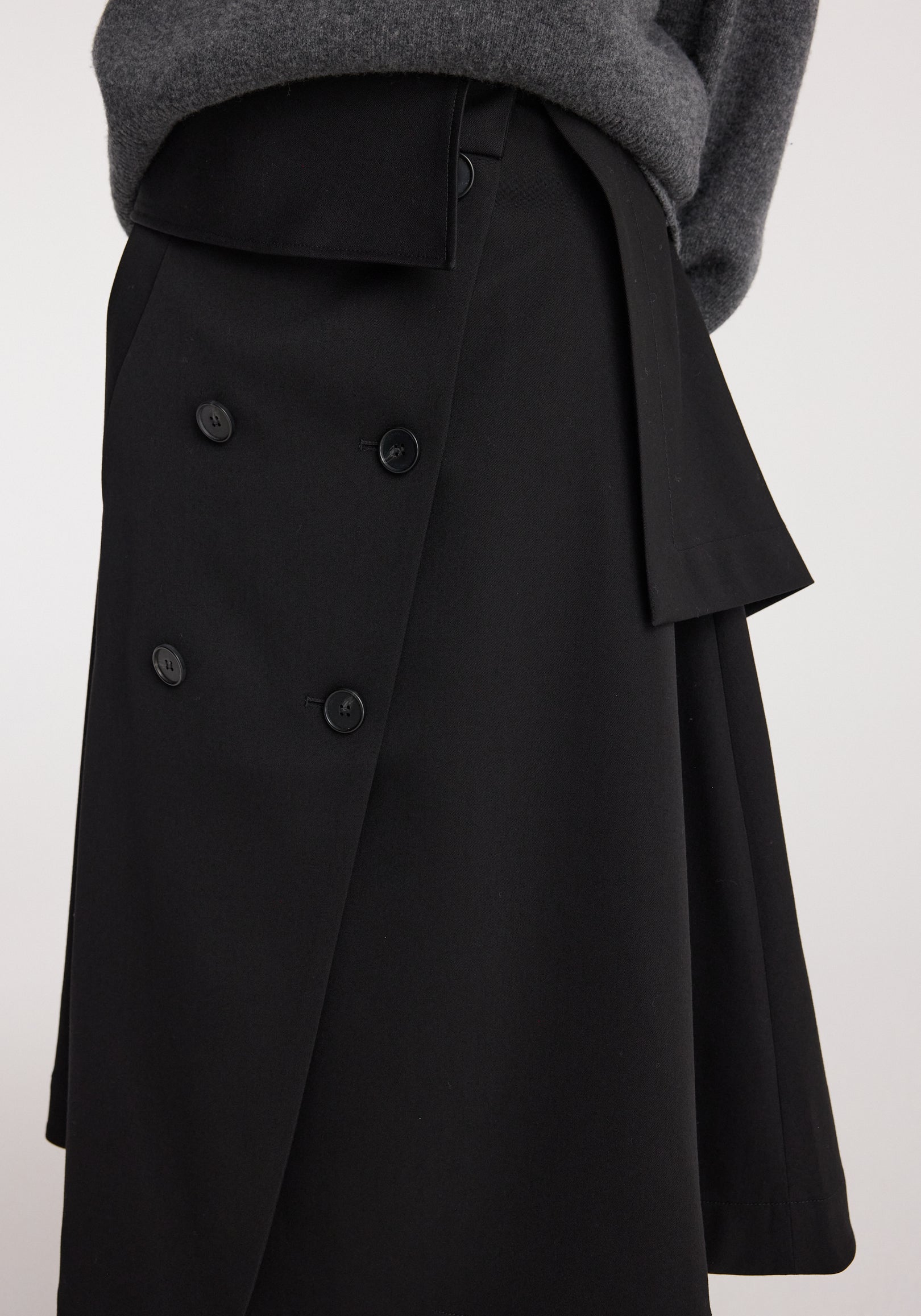Close-up of a black Deconstructed trench skirt | black featuring button details and an asymmetrical layered design paired with a dark grey sweater.