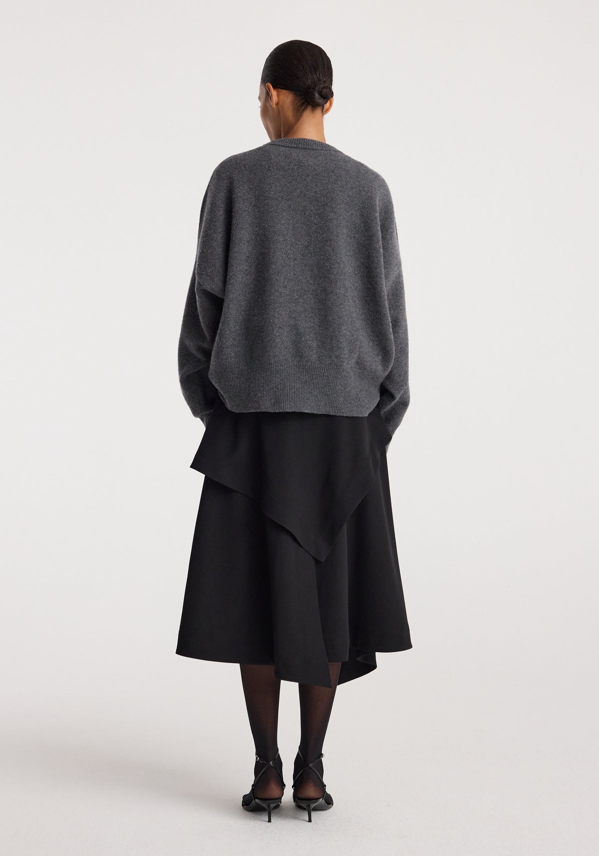 Woman wearing black layered asymmetric skirt with an oversized dark gray sweater and black heels, showcasing the back view of the Deconstructed trench skirt | black.