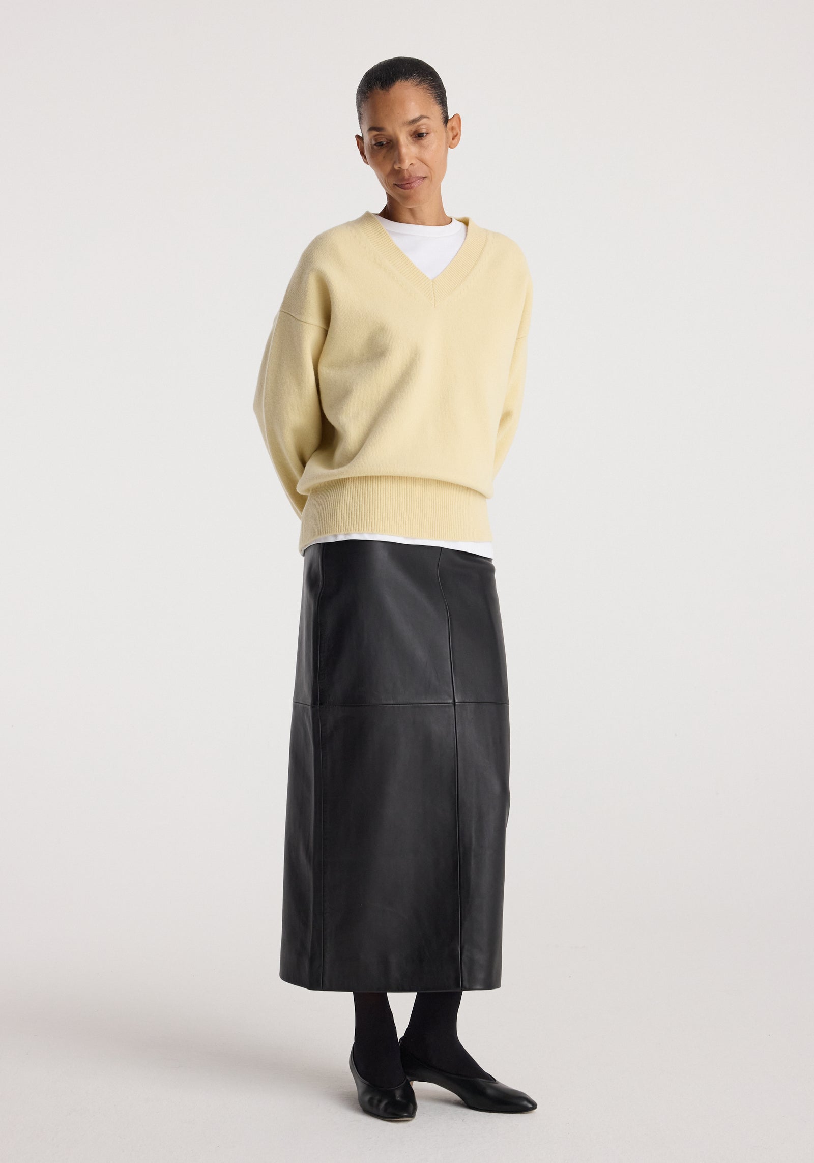 Woman wearing an elongated leather skirt | black paired with a cream V-neck sweater and black shoes, standing against a plain white background.