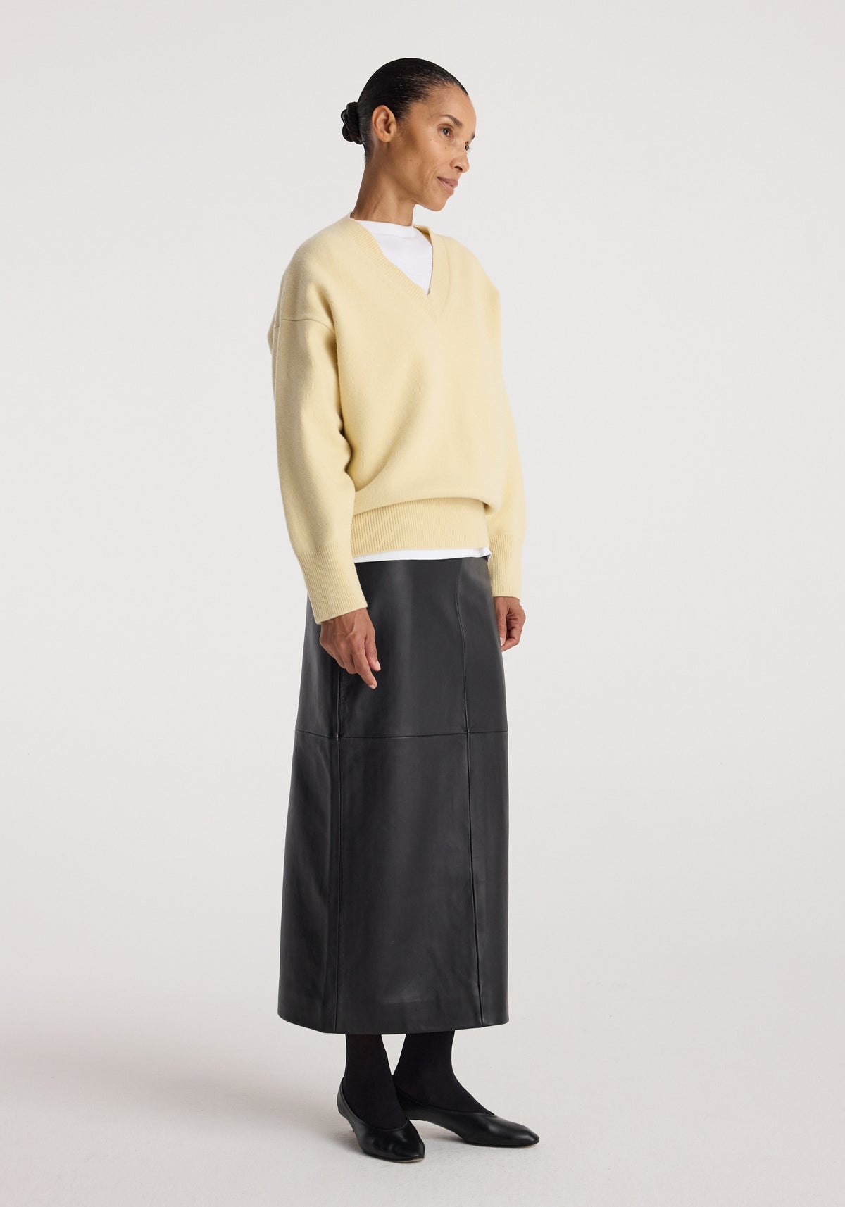 Woman wearing an elongated leather skirt | black paired with a cream sweater, black tights, and black flat shoes, standing against a plain white background.