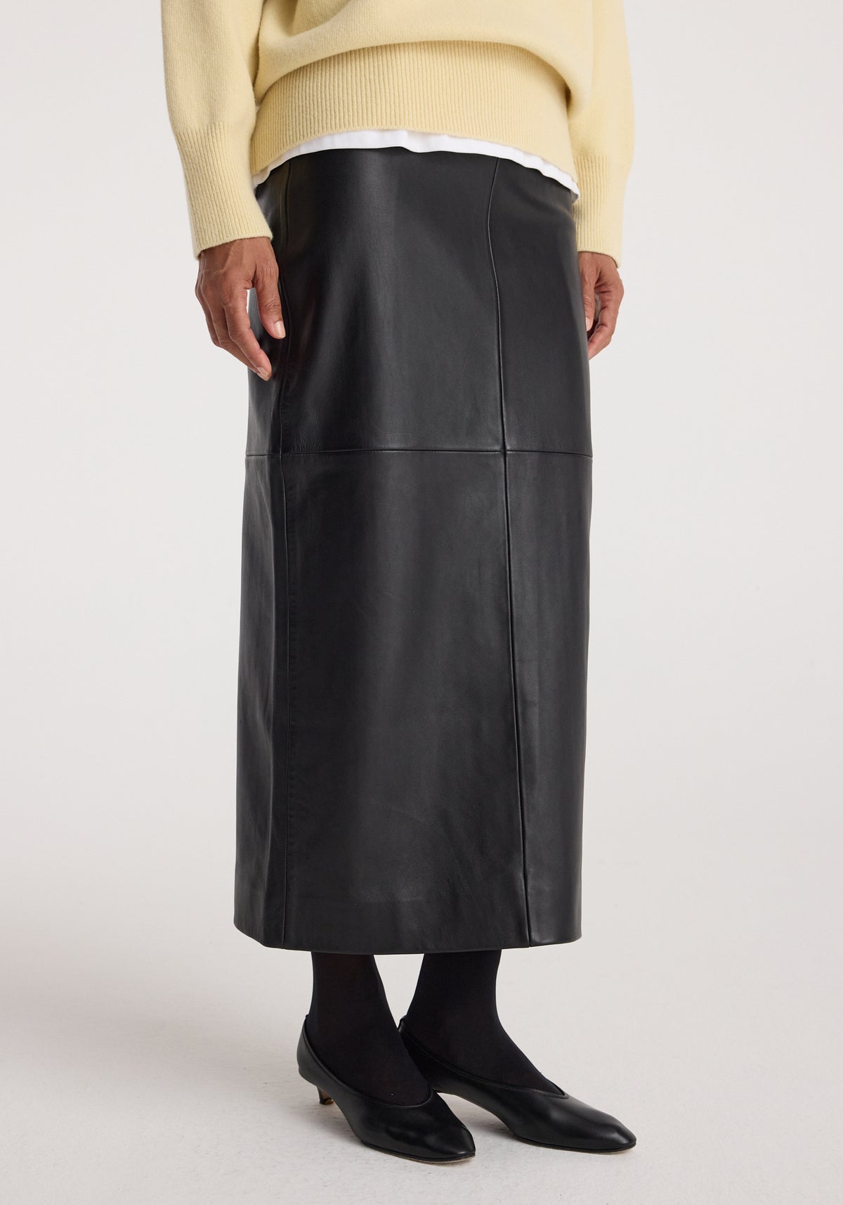 Elongated leather skirt | black worn with black tights and black low-heeled shoes, paired with a cream sweater layered over a white shirt.
