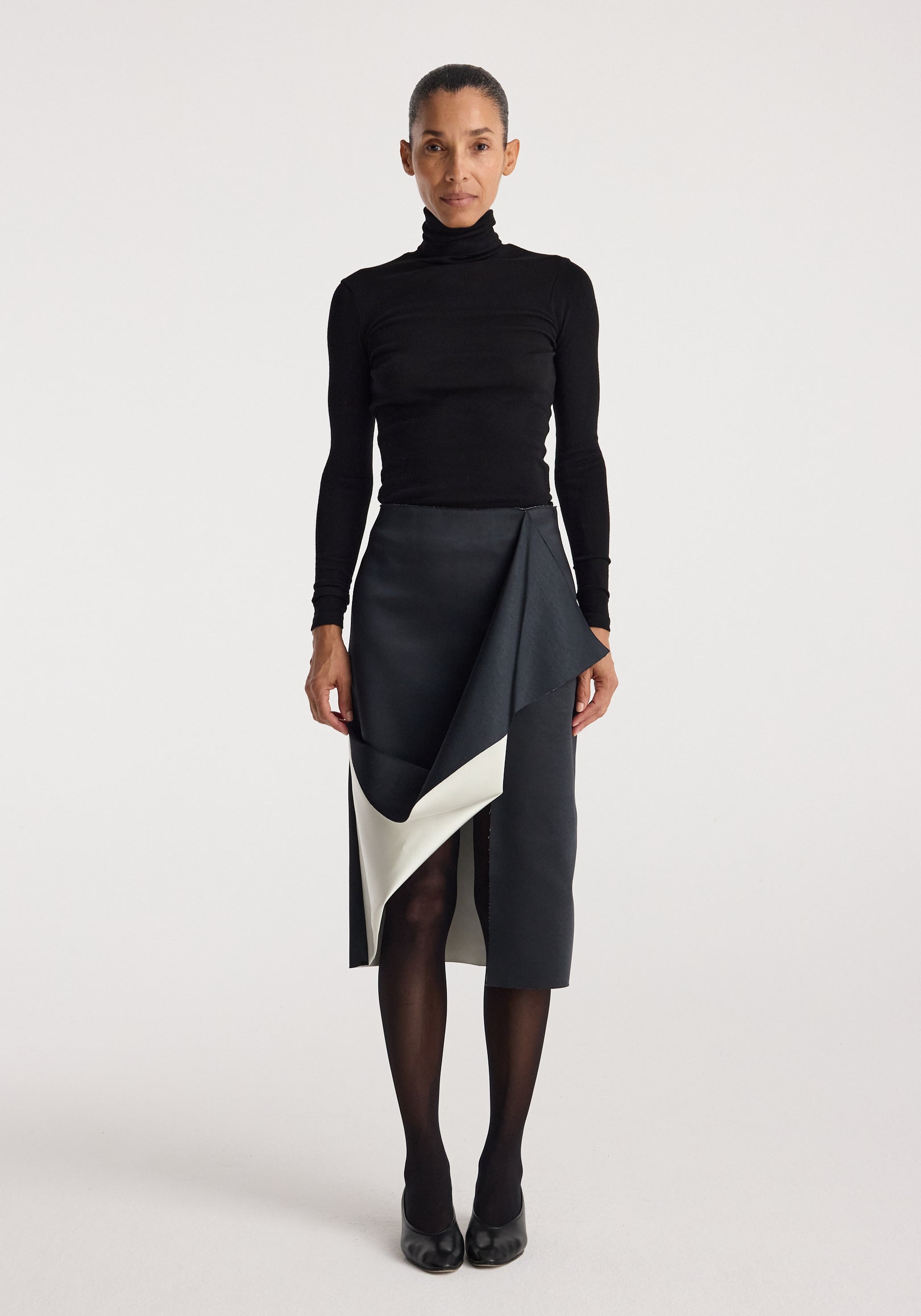 A woman wearing a black turtleneck top paired with a Sculptural double-satin skirt | black/white featuring a draped, asymmetrical design and contrasting white lining, styled with black tights and black heels.