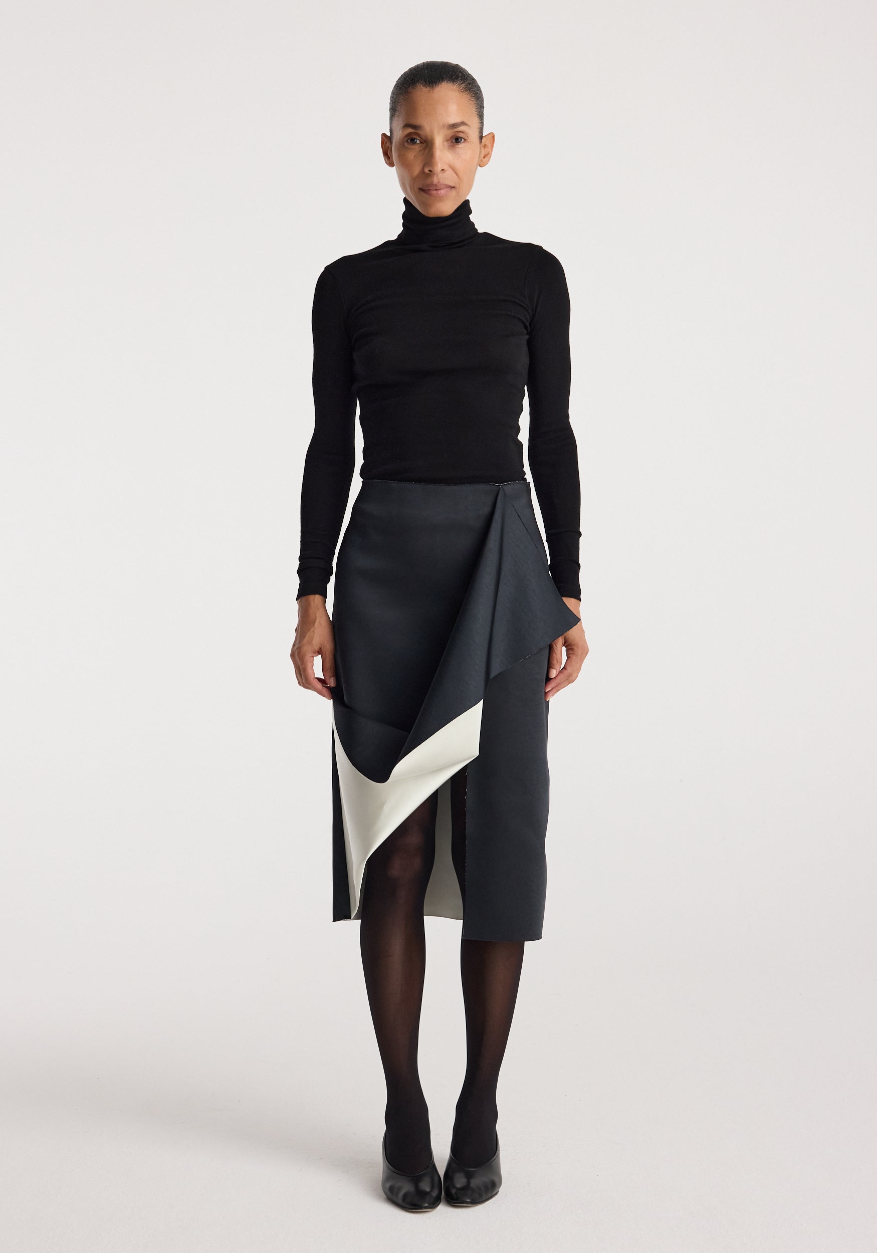 A woman wearing a black turtleneck top paired with a Sculptural double-satin skirt | black/white featuring a draped, asymmetrical design and contrasting white lining, styled with black tights and black heels.