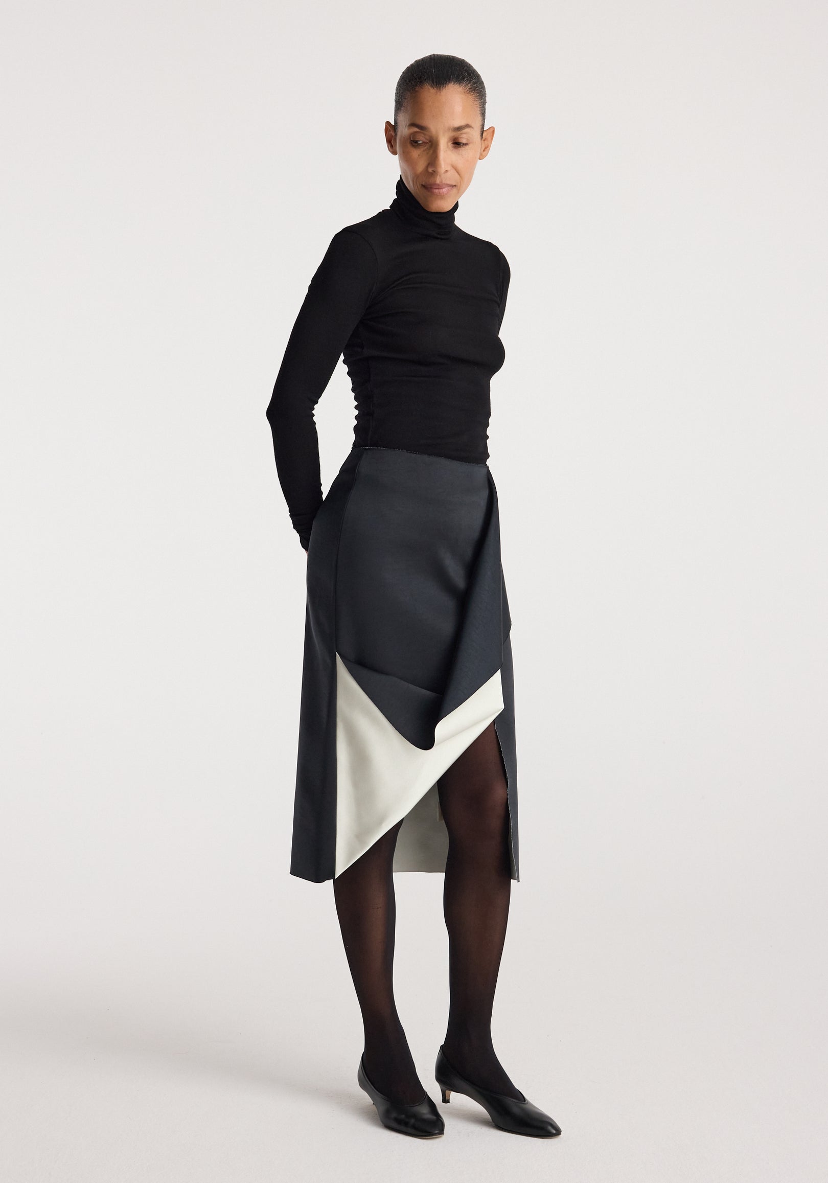 Woman wearing a black long-sleeve top paired with a Sculptural double-satin skirt | black/white featuring a folded detail and color contrast, styled with black tights and shoes.
