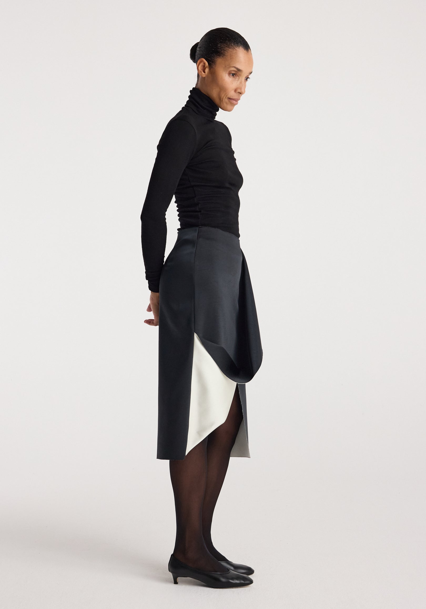 Woman wearing a black turtleneck paired with a sculptural double-satin skirt | black/white featuring a draped overlay and contrasting inner panel, styled with black tights and low heels.