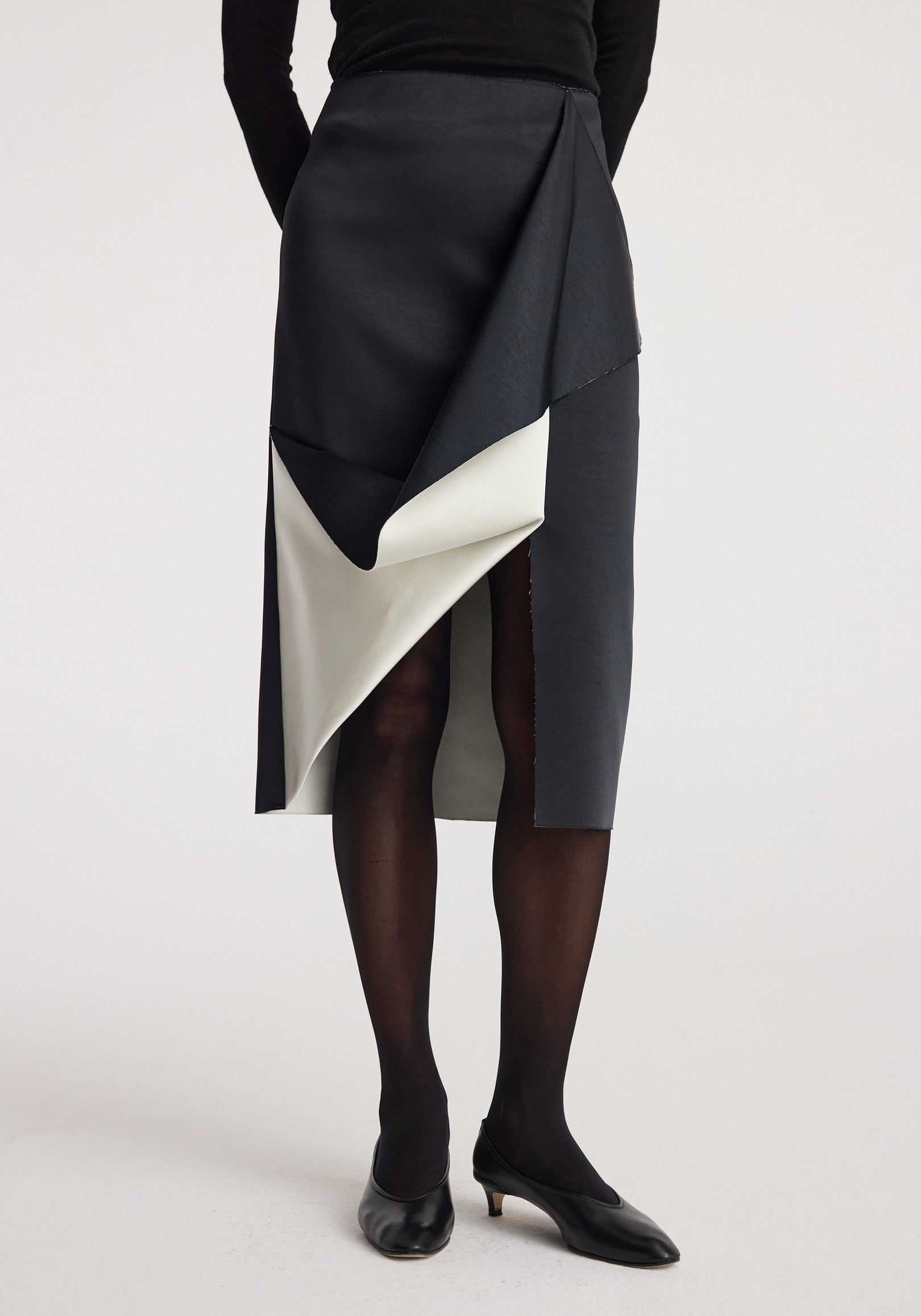 Woman wearing a sculptural double-satin skirt | black/white featuring layered contrasting black and white panels, paired with black tights and black kitten heel shoes.