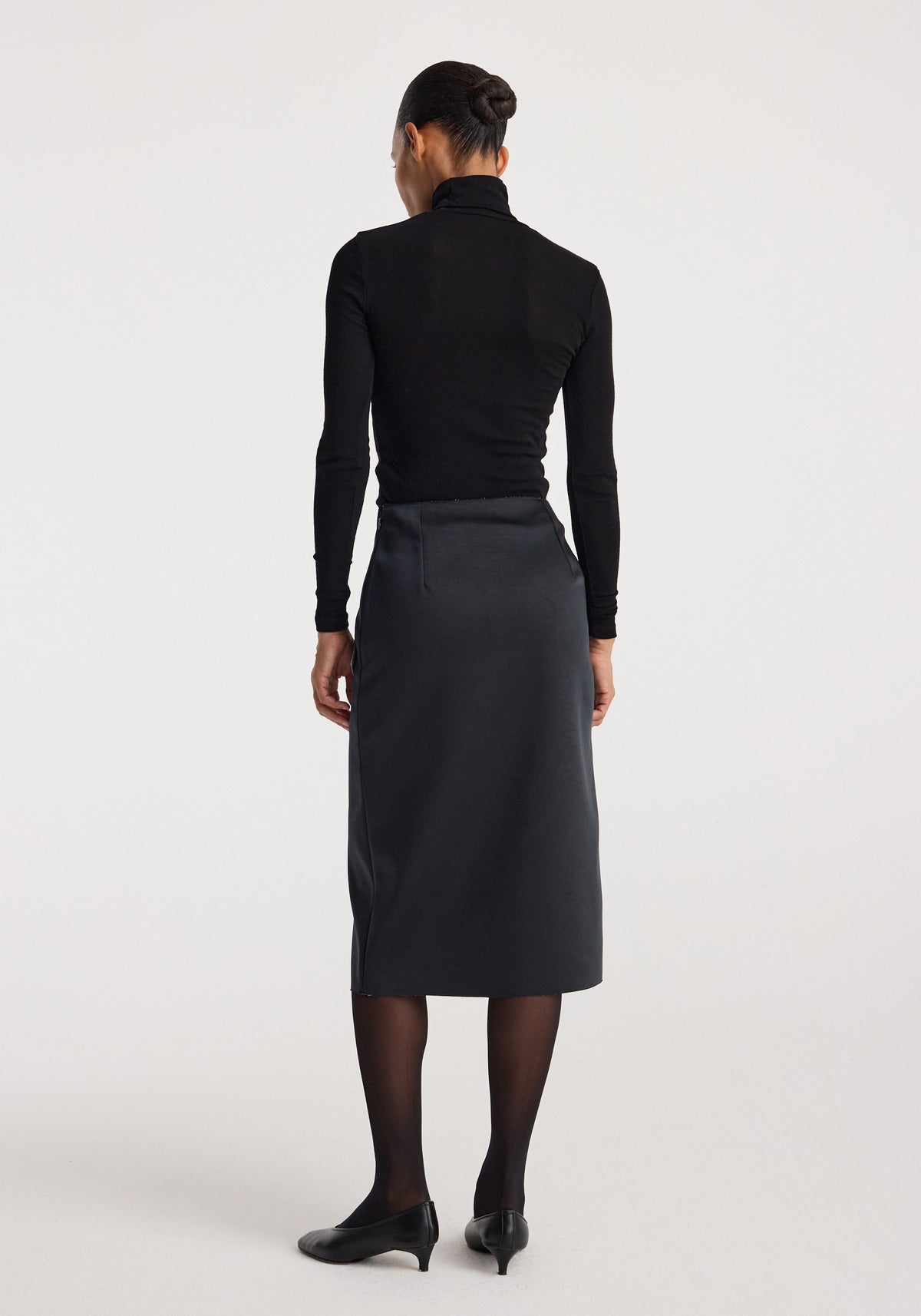 Woman wearing a Sculptural double-satin skirt | black/white, shown from behind, styled with black sheer tights, black pointed shoes, and a black long-sleeve fitted top.