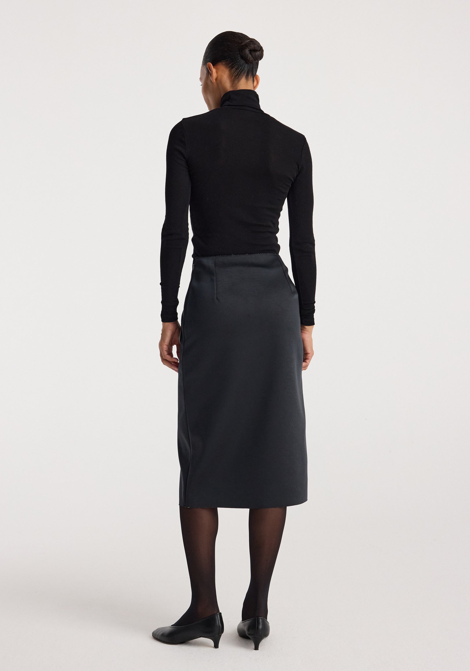 Woman wearing a Sculptural double-satin skirt | black/white, shown from behind, styled with black sheer tights, black pointed shoes, and a black long-sleeve fitted top.