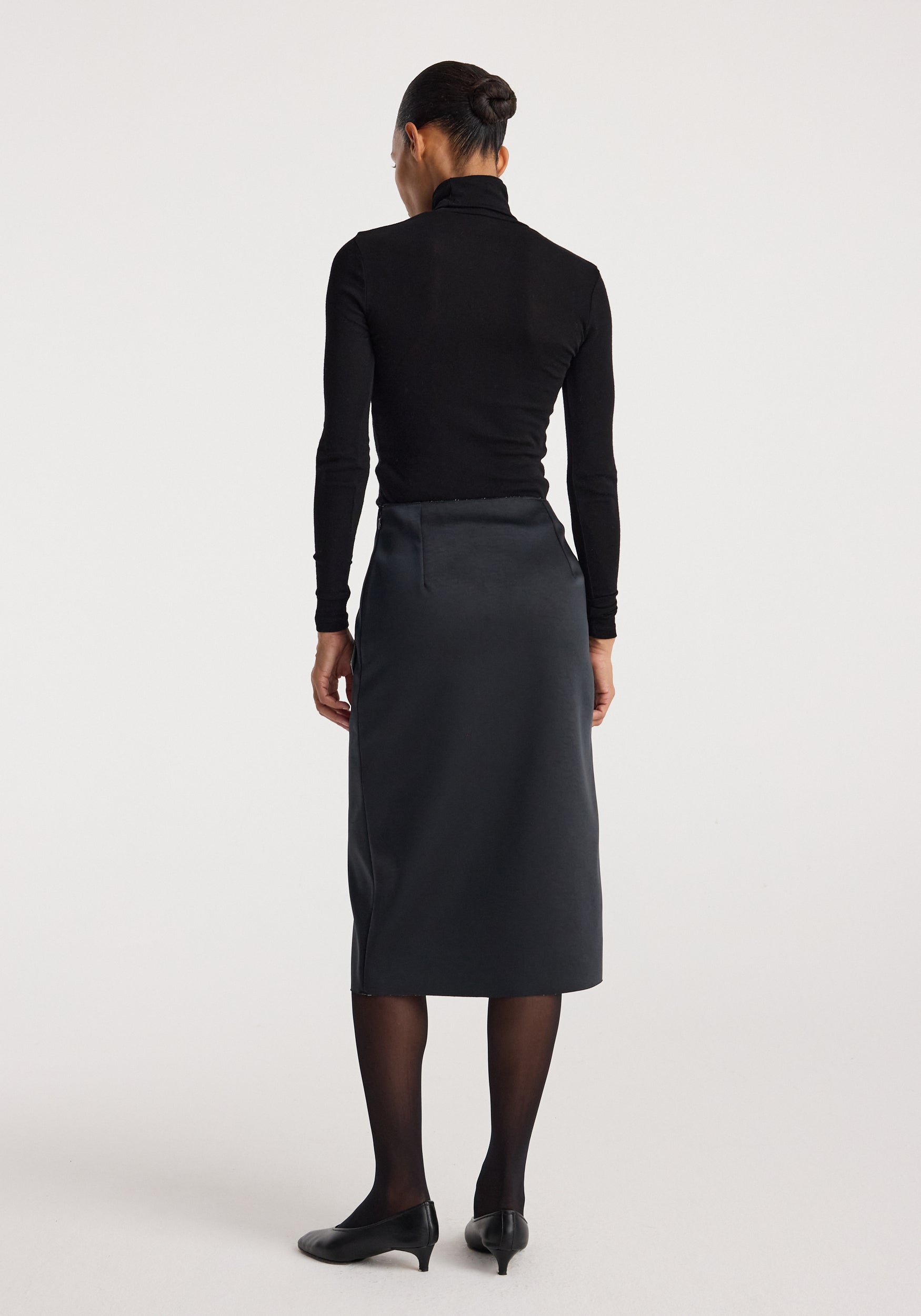 Woman wearing a Sculptural double-satin skirt | black/white, shown from behind, styled with black sheer tights, black pointed shoes, and a black long-sleeve fitted top.
