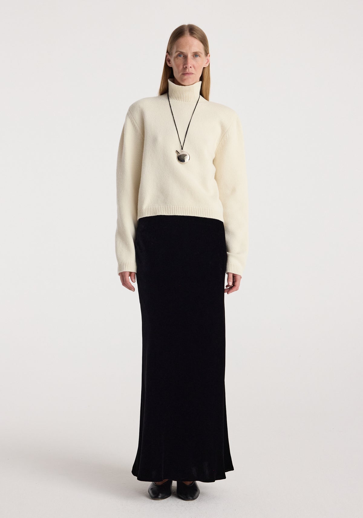 Woman wearing a cream turtleneck sweater with a long black fluid velvet skirt | black, paired with black shoes and a silver pendant necklace.