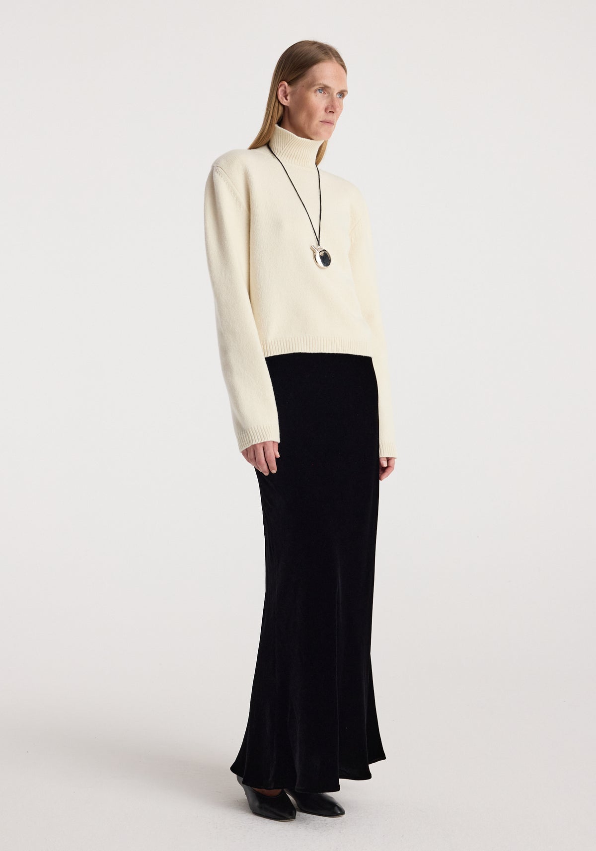Woman wearing a cream turtleneck sweater paired with a black floor-length Fluid velvet skirt | black and black pointed shoes standing against a plain background.