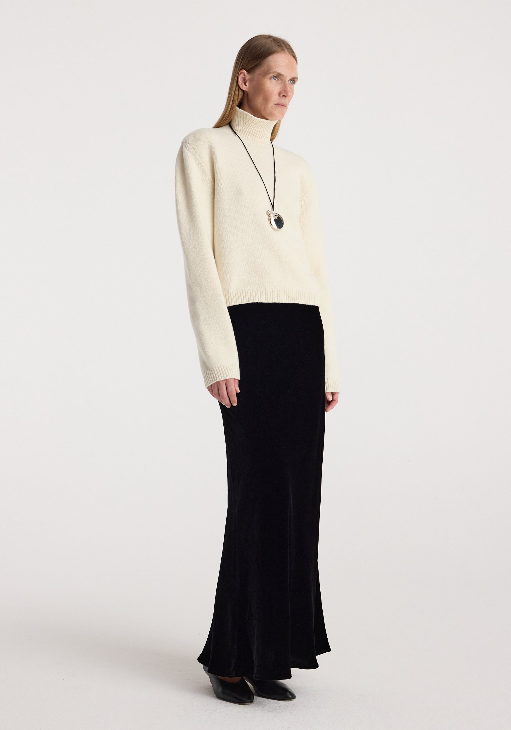 Woman wearing a cream turtleneck sweater paired with a black floor-length Fluid velvet skirt | black and black pointed shoes standing against a plain background.