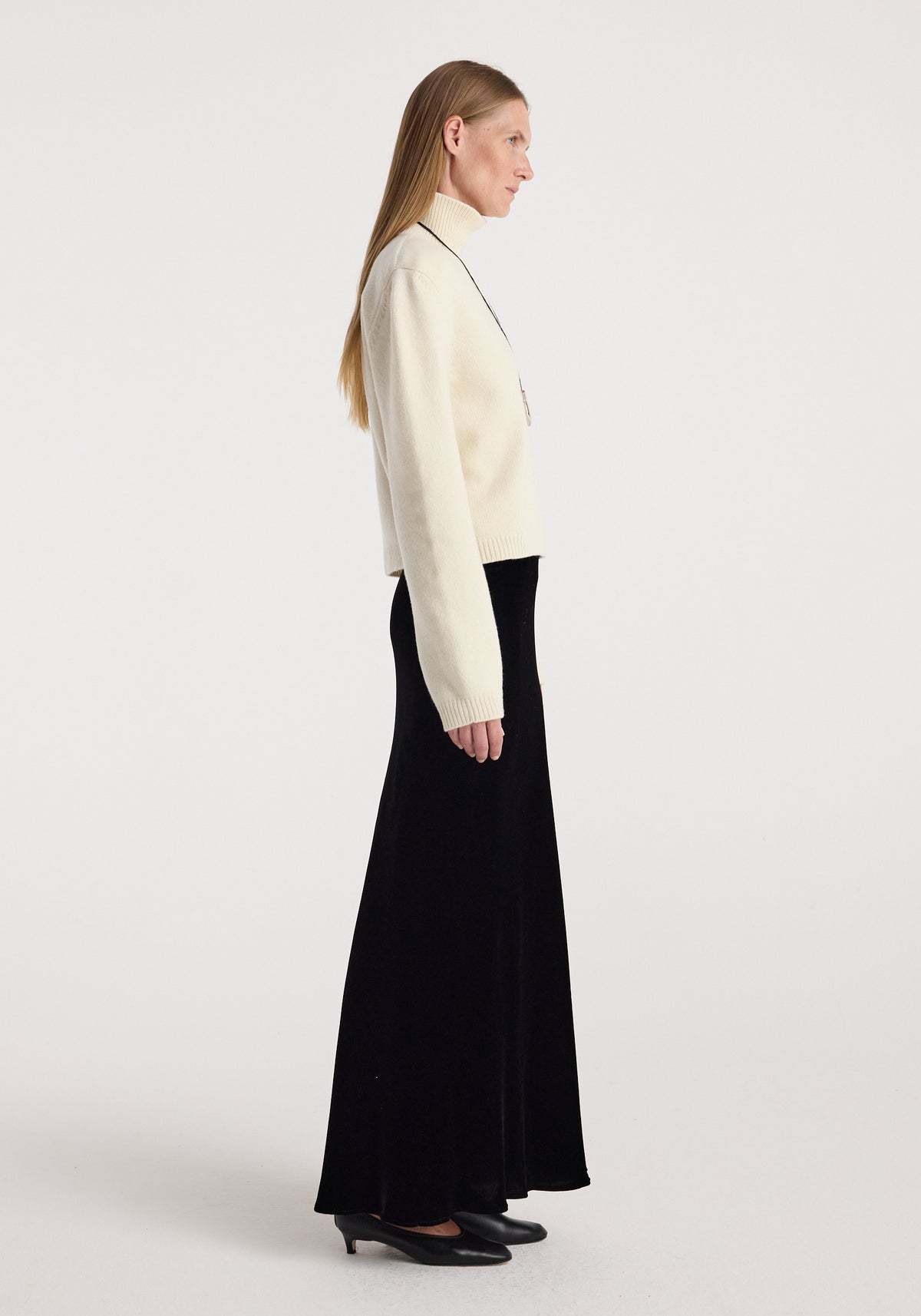 Woman wearing a cream turtleneck sweater and a long black Fluid velvet skirt | black, standing in profile against a plain white background with black low-heeled shoes.