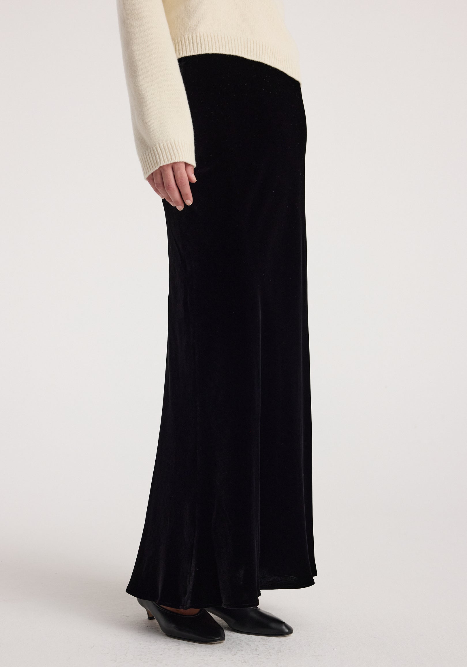 Side view of a person wearing a long black Fluid velvet skirt | black paired with black shoes and a cream sweater sleeve visible