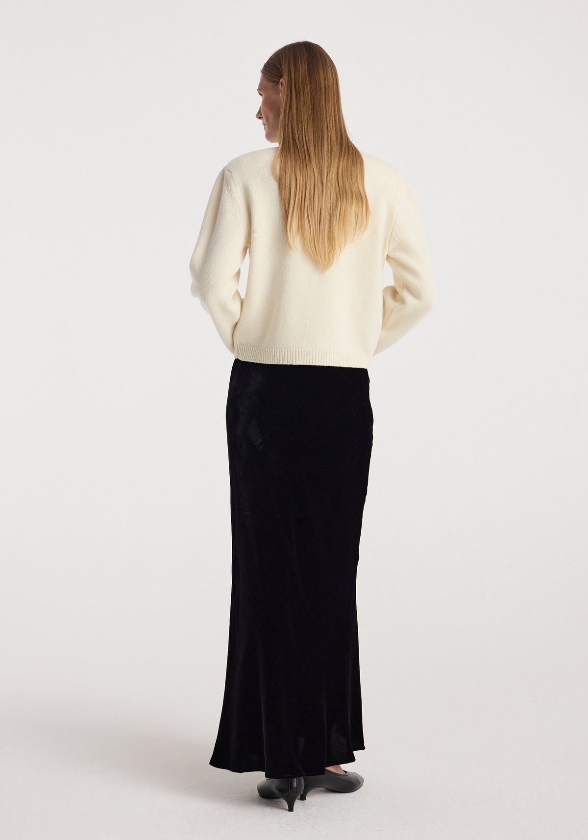 Woman seen from behind wearing a long black velvet skirt paired with a cream-colored sweater and black heels, showcasing the Fluid velvet skirt | black.