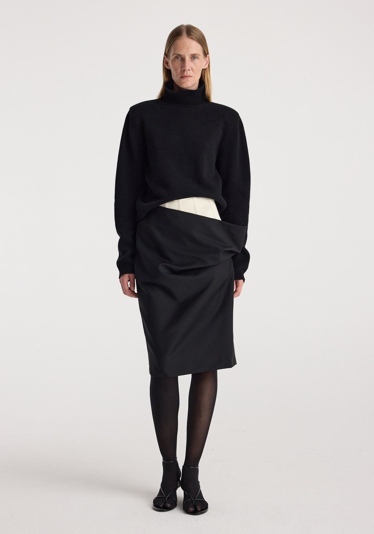 Woman wearing a black turtleneck sweater paired with a Double waistband skirt | black, sheer black tights, and black open-toe heels, standing against a plain white background.