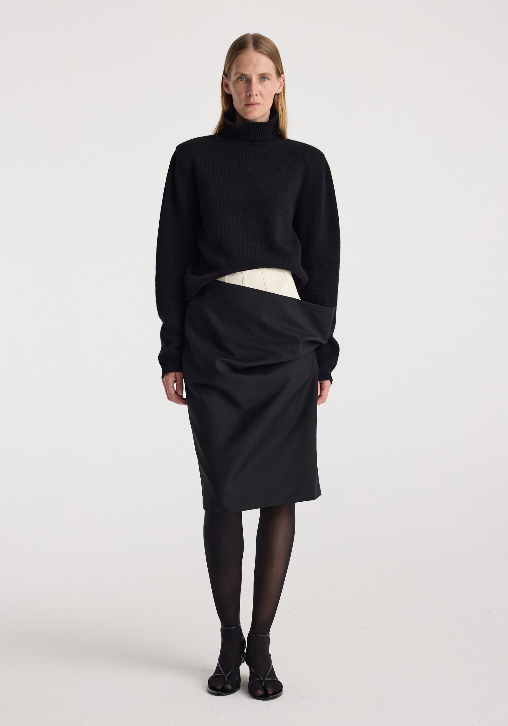 Woman wearing a black turtleneck sweater paired with a Double waistband skirt | black, sheer black tights, and black open-toe heels, standing against a plain white background.