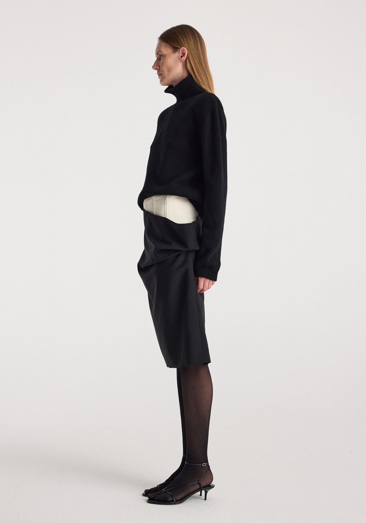 Profile view of a woman wearing a black turtleneck sweater paired with a Double waistband skirt | black, sheer black tights, and black kitten heel shoes against a plain background.