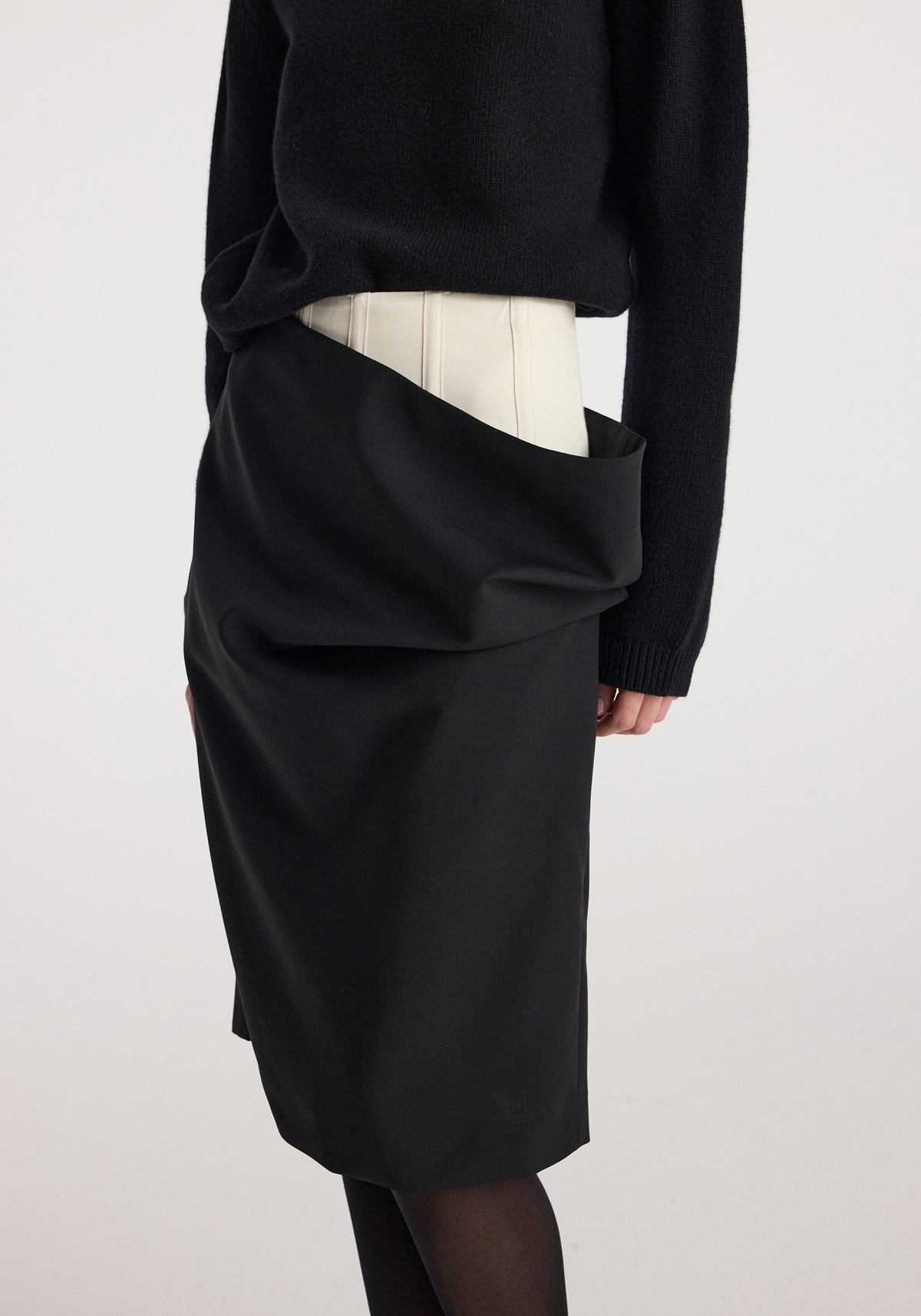 Model wearing a black double waistband skirt | black with a draped upper layer over a fitted underlayer, paired with a black sweater and black tights.