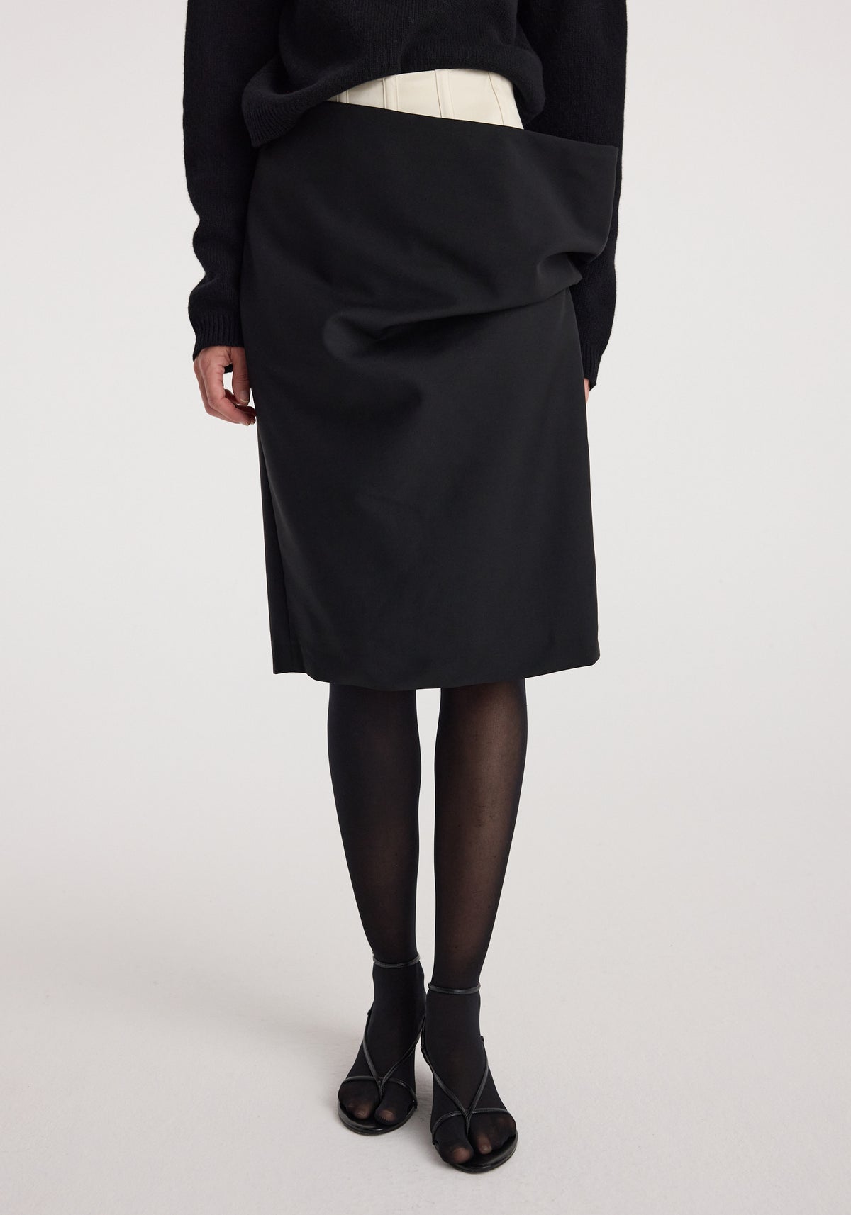 Woman wearing a black double waistband skirt | black paired with black tights and open-toe high heel shoes, shown from the waist down against a neutral background.