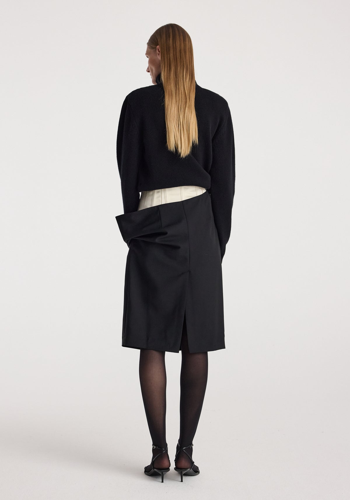 Woman wearing a black double waistband skirt | black with a white layer visible at the waist, paired with black sheer tights, black heels, and a black long-sleeve top, shown from the back.