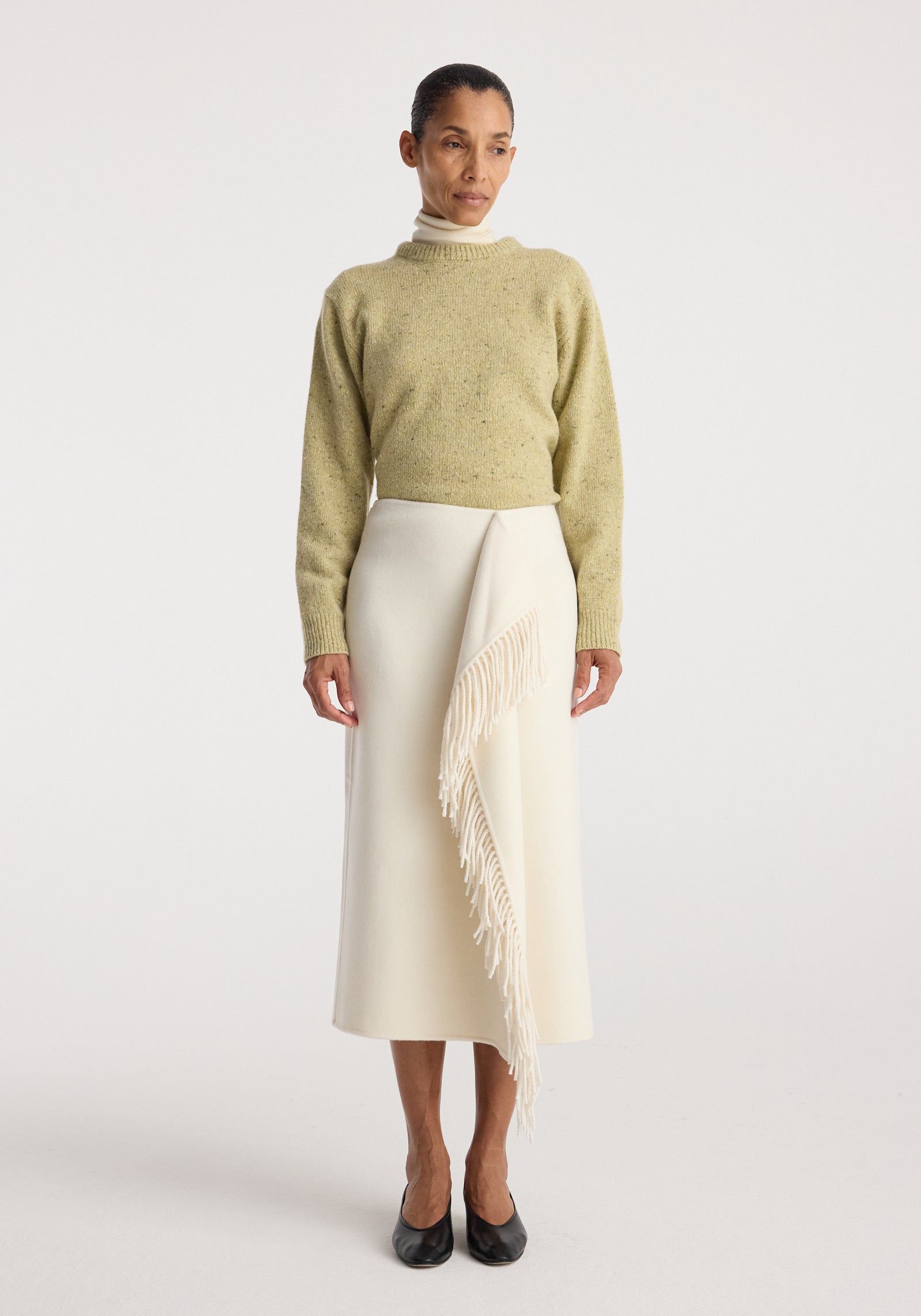 Woman wearing a beige sweater and black heels posing in a minimalist studio, featuring the Double-faced fringe skirt | off white with asymmetrical fringe detail on the front.
