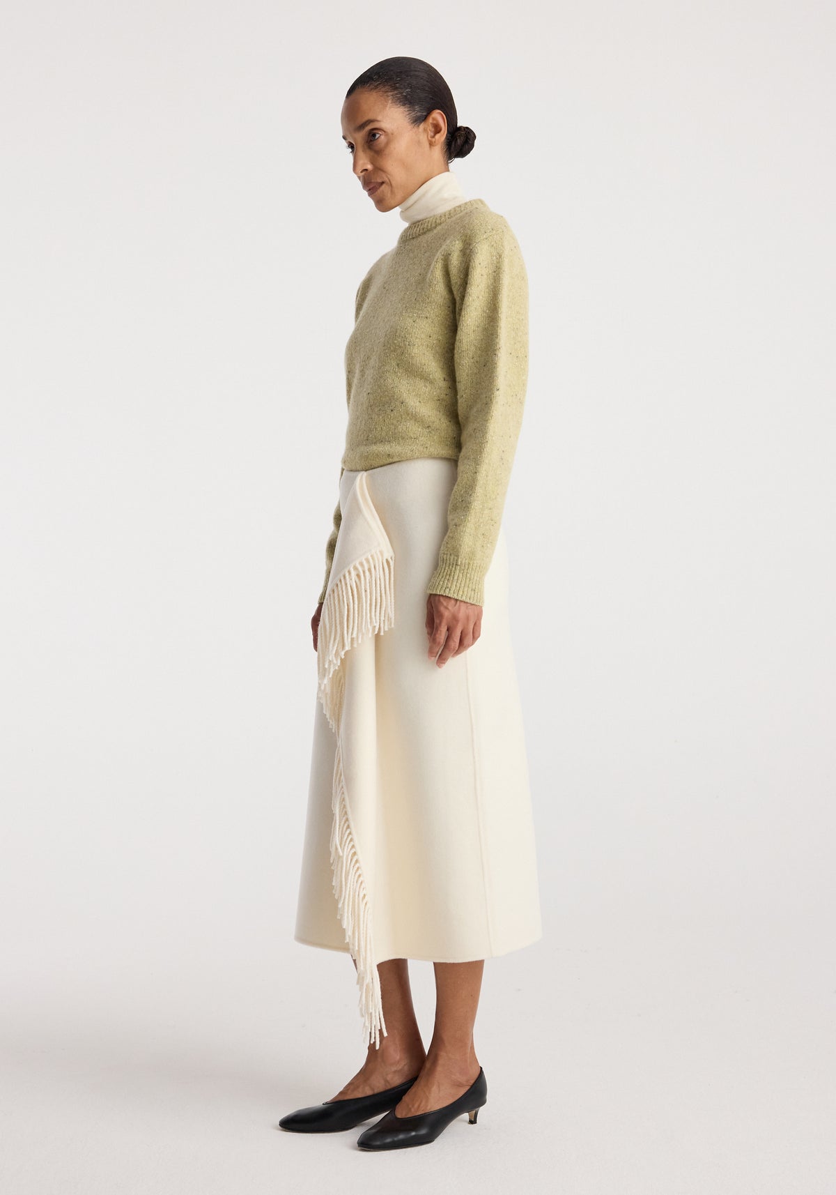 Woman wearing a soft green sweater and a cream-colored double-faced fringe skirt | off white with long flowing fringe detail paired with black low-heeled shoes.