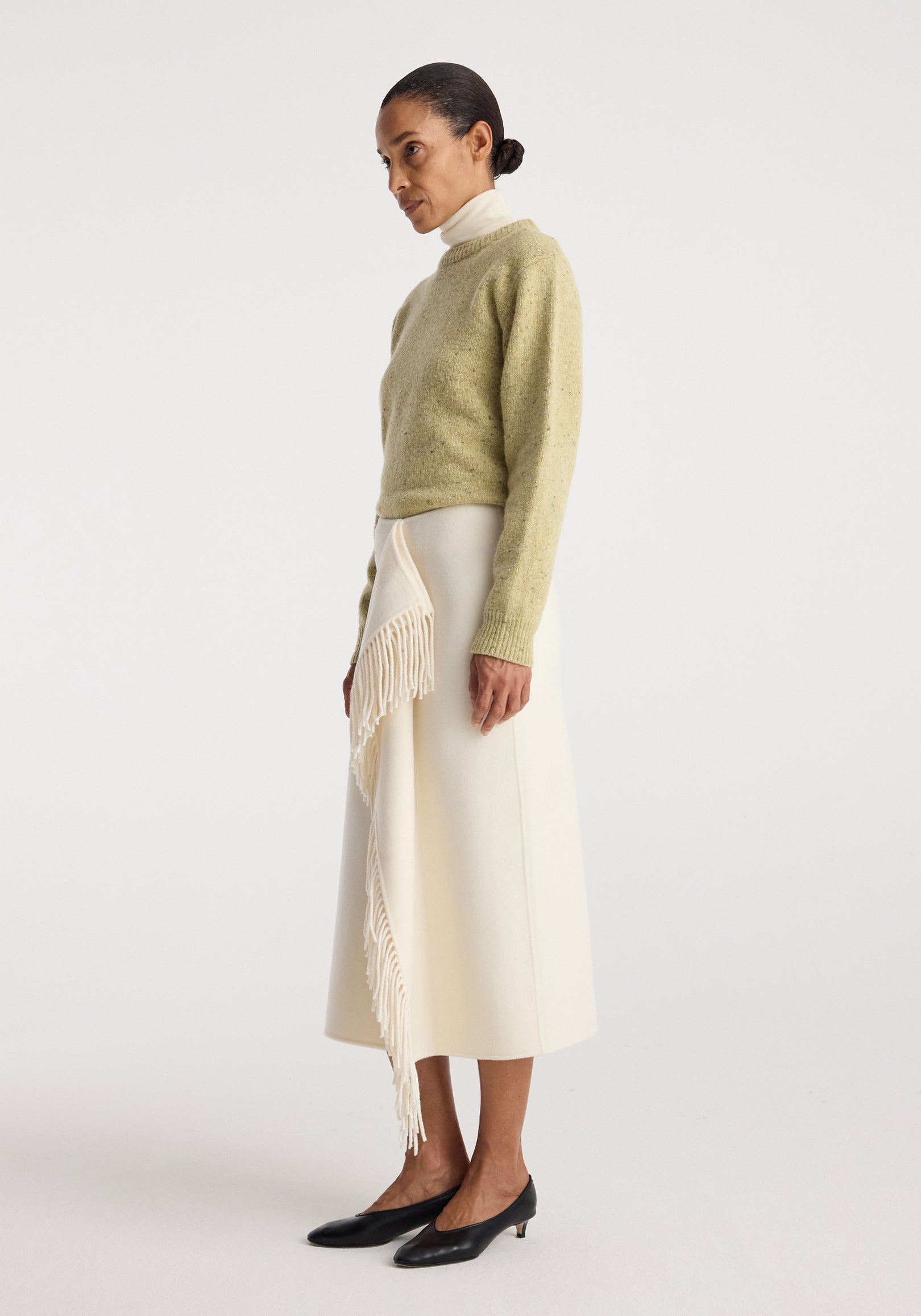 Woman wearing a soft green sweater and a cream-colored double-faced fringe skirt | off white with long flowing fringe detail paired with black low-heeled shoes.
