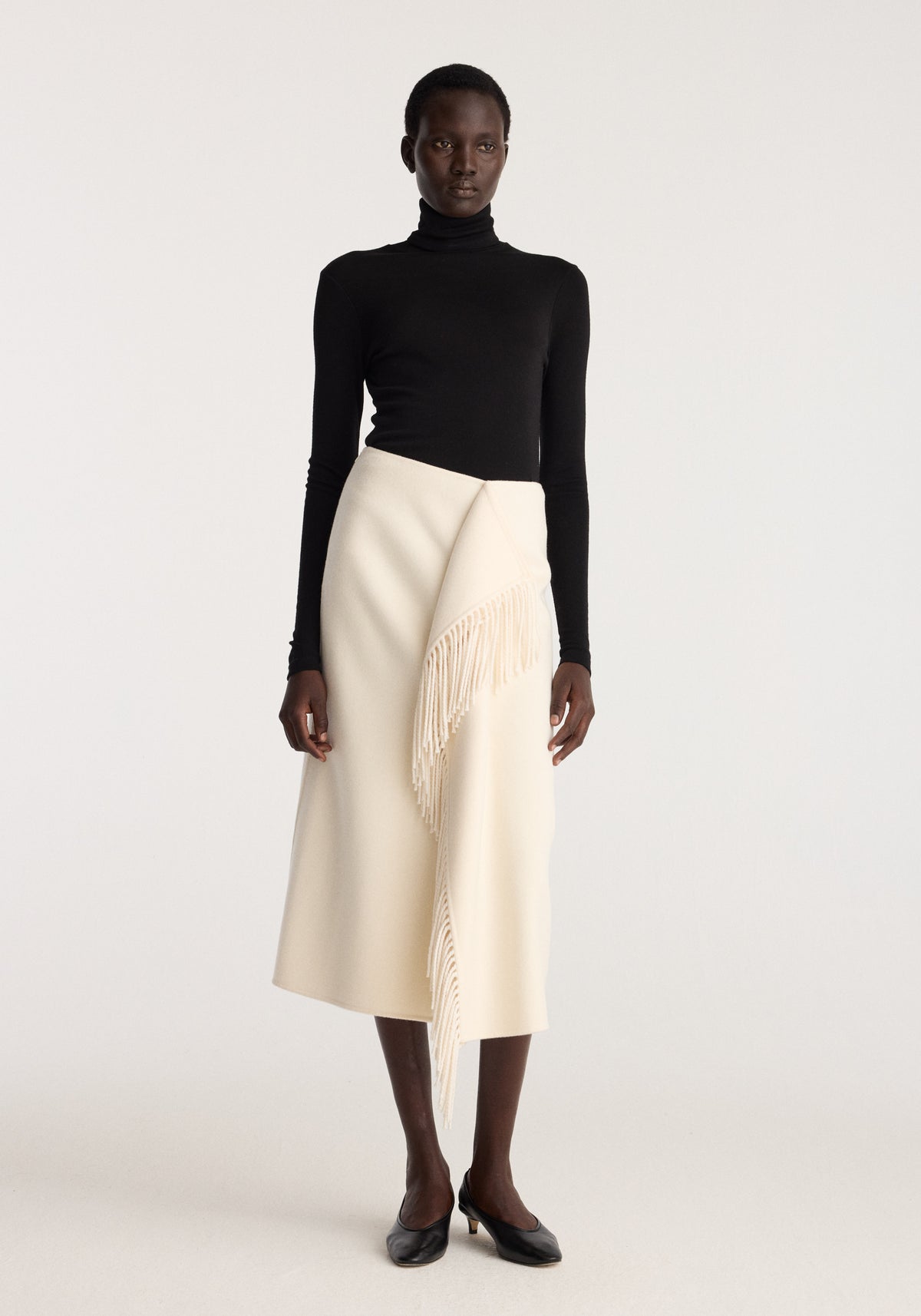 Double-faced fringe skirt | off white worn with a black turtleneck showing an asymmetrical wrap front and long fringe detail on a model in studio lighting