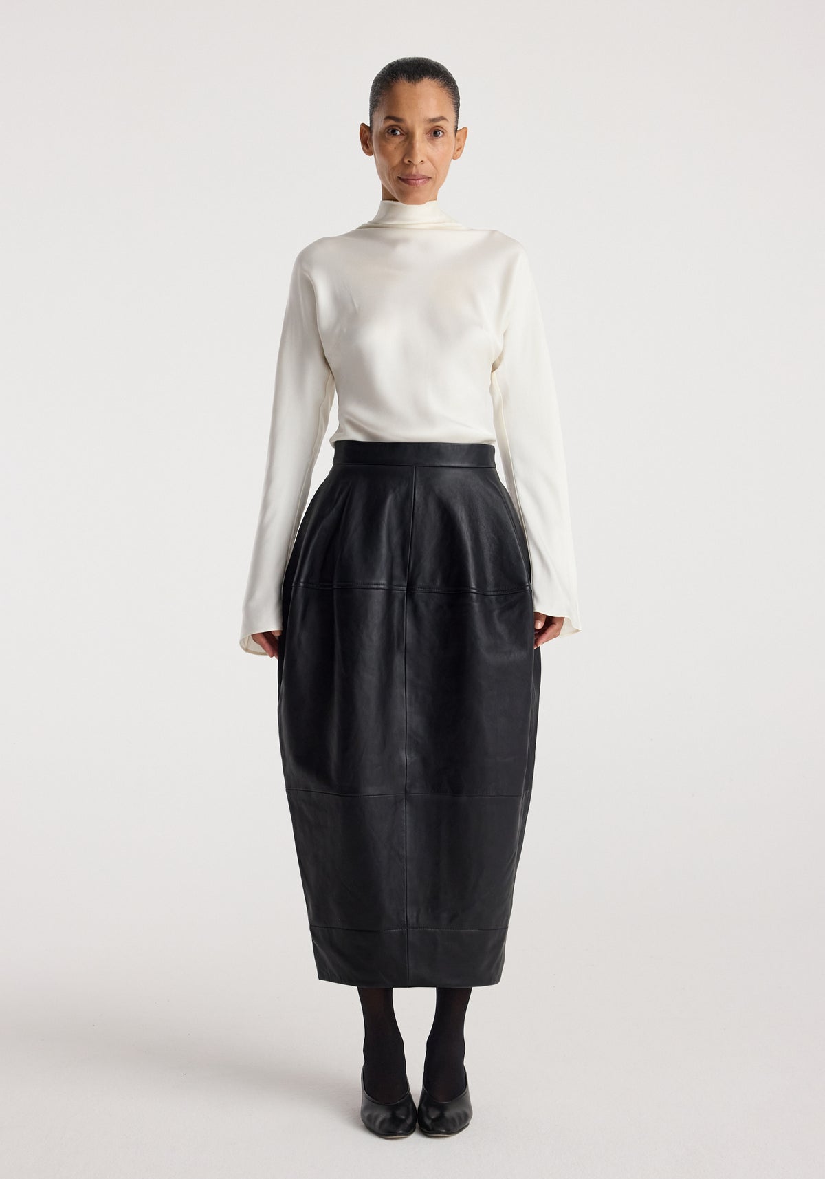 Woman wearing a white long-sleeve top paired with a black Sculptural leather skirt | black, standing against a plain white background.