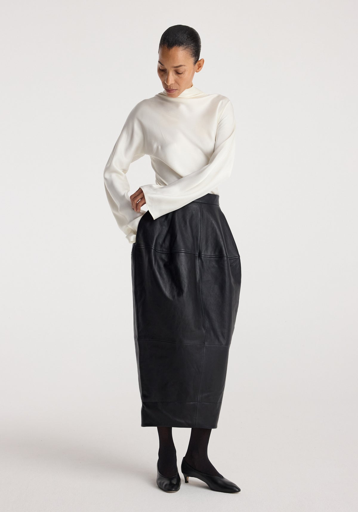 Woman wearing a high-waisted black Sculptural leather skirt | black paired with a white long-sleeve top and black heels against a plain background.