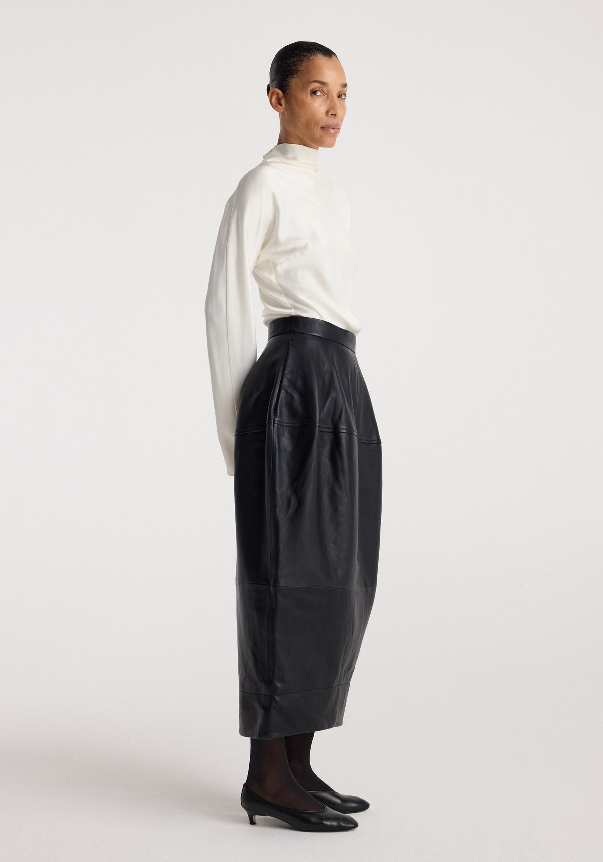 Woman wearing a white high-neck blouse and black Sculptural leather skirt | black with a midi length and detailed stitching, paired with black tights and low-heeled shoes.