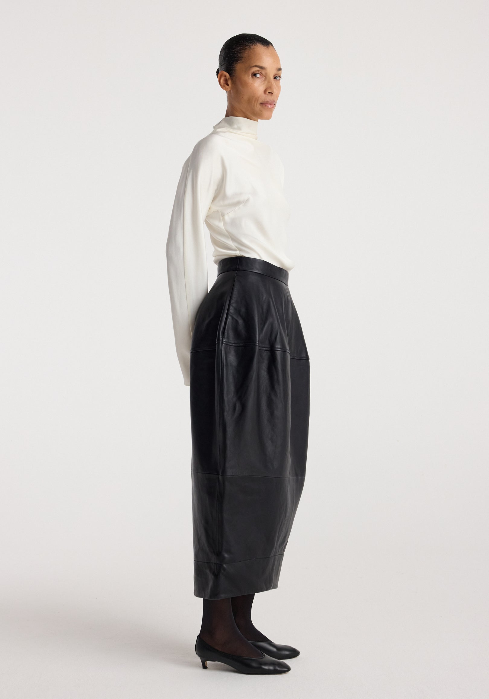 Woman wearing a white high-neck blouse and black Sculptural leather skirt | black with a midi length and detailed stitching, paired with black tights and low-heeled shoes.