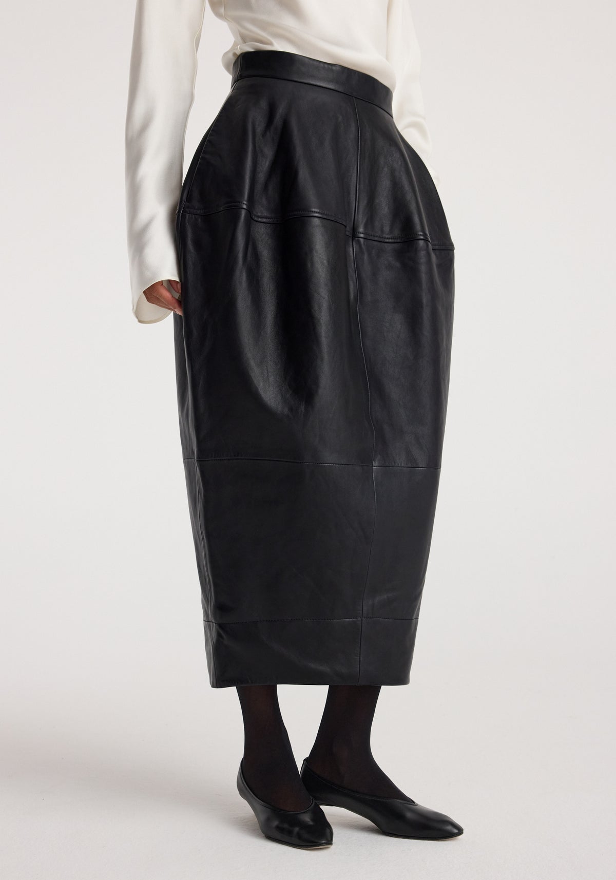 Woman wearing a sculptural leather skirt | black with a high waist, midi length, and subtle texture, paired with black tights and black flats.