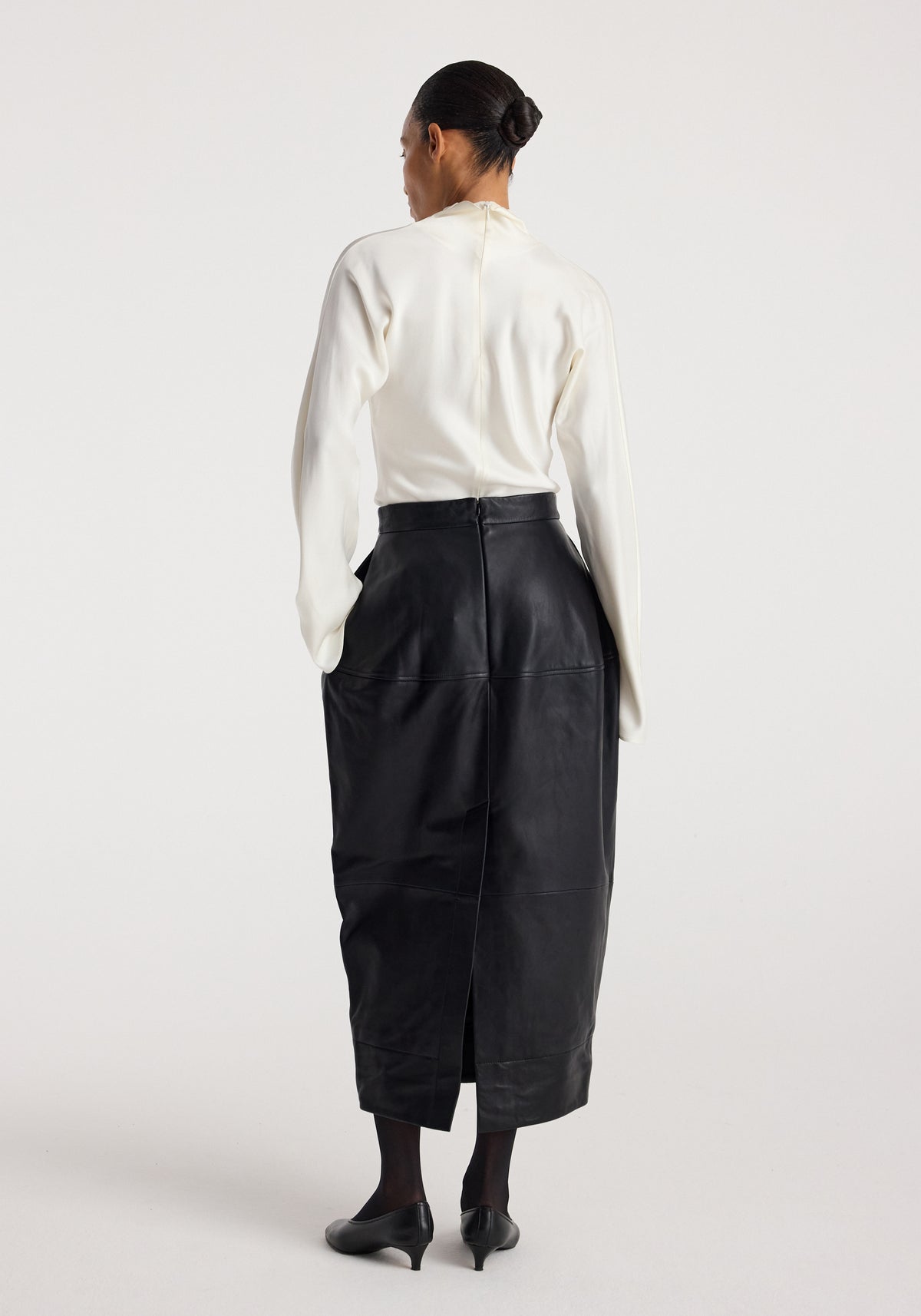 Woman wearing a Sculptural leather skirt | black styled with a white long-sleeve top, standing facing away, showing the back details and slit of the skirt.