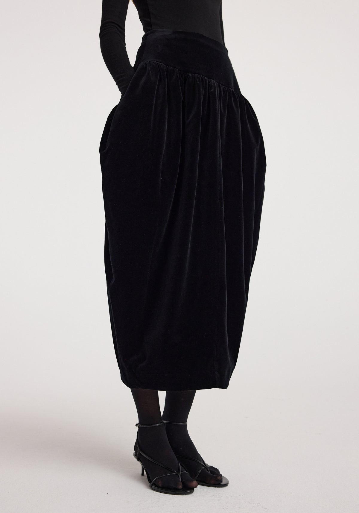 Sculptural volume velvet skirt | black worn by a model, featuring a high waist, gathered details, voluminous midi silhouette, and black strappy heels