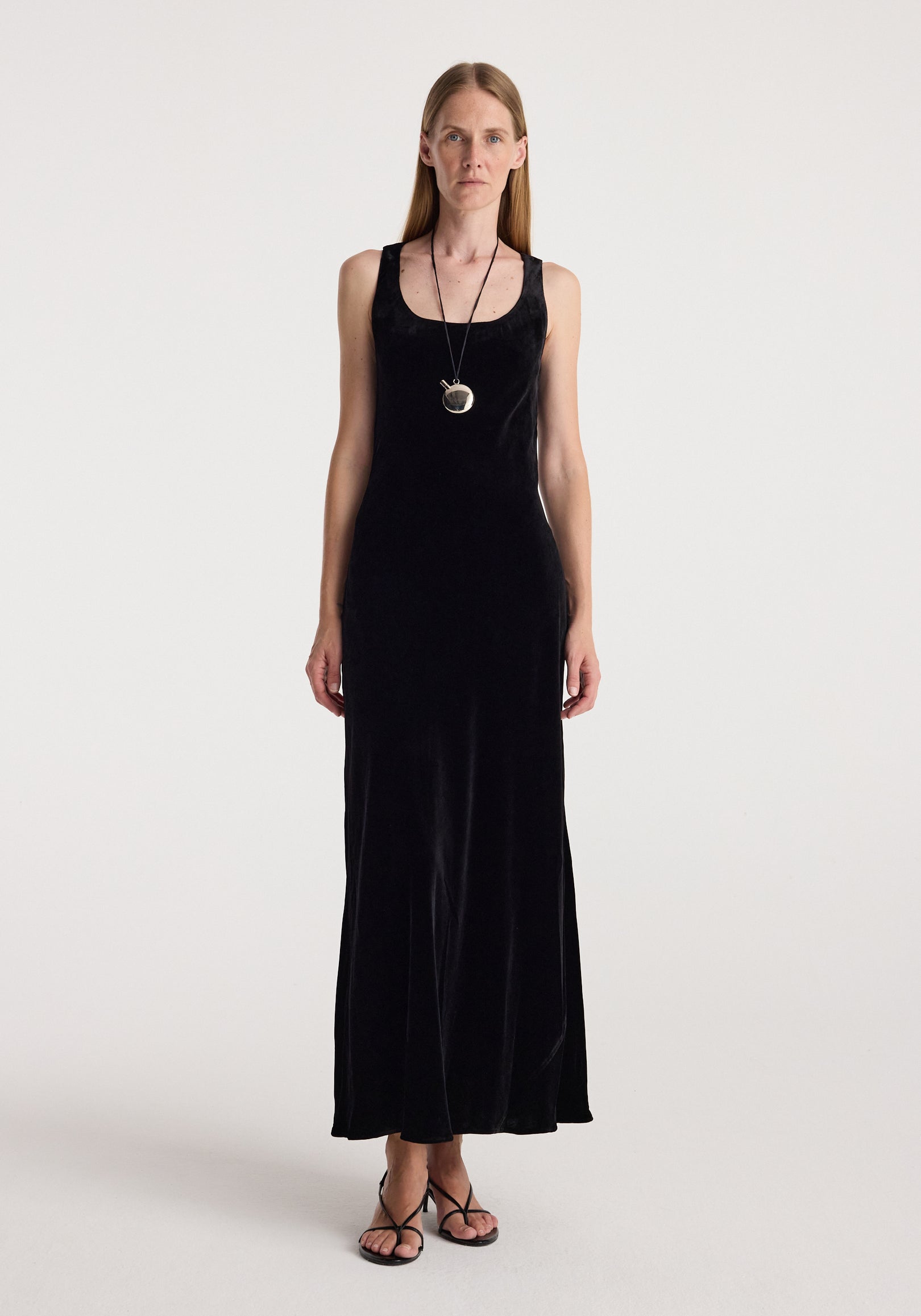 Woman wearing a long black velvet dress with a scoop neckline, paired with a pendant necklace and strappy sandals, standing against a plain light background, Scoop-neck velvet dress | black.